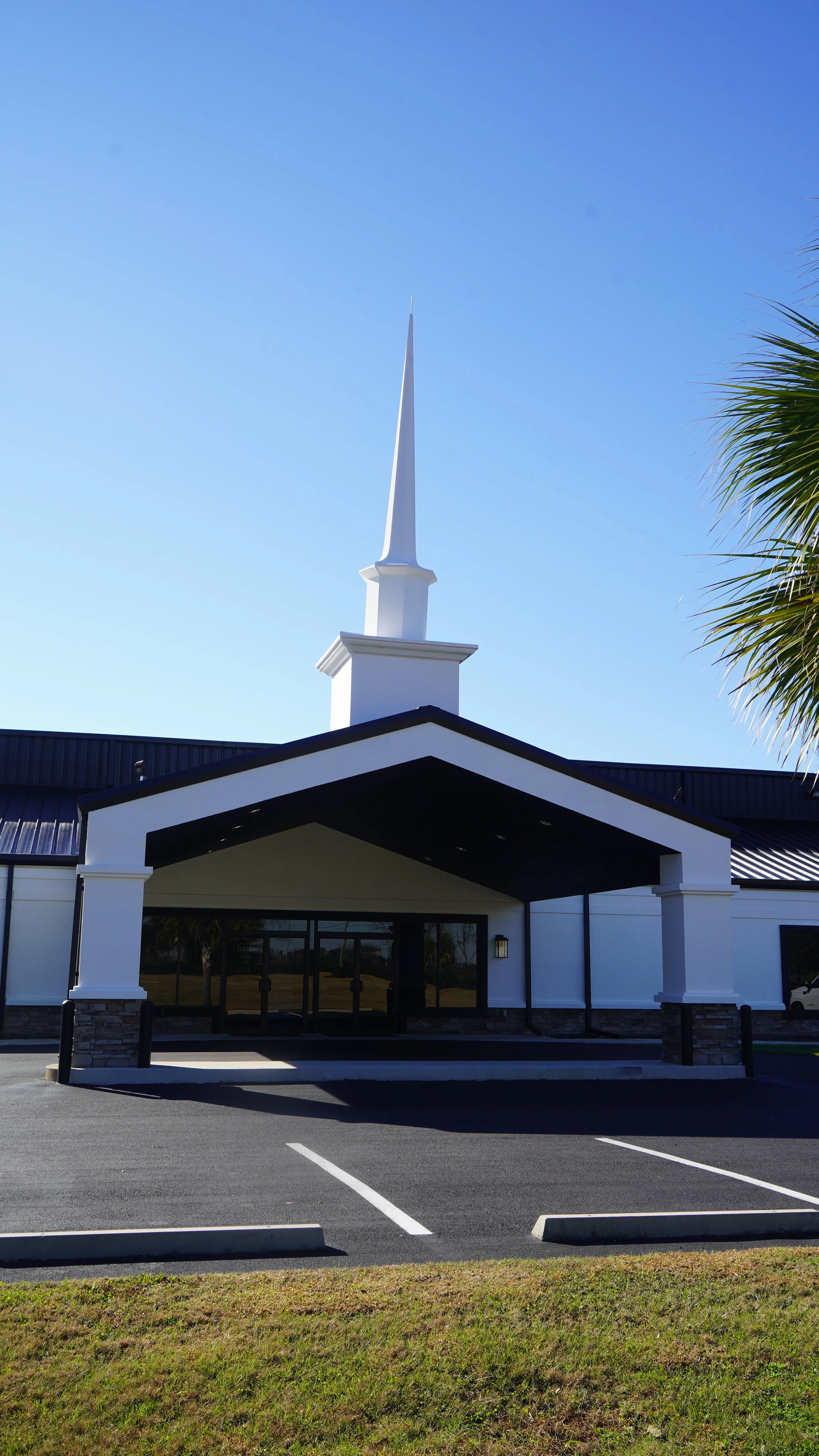 Front view of a church building with a tall white steeple, black roof, and a covered entrance. There is a parking lot in front of the building with parking spaces and grass on the sides. A palm tree is visible on the right side of the image, and the sky is clear and blue.