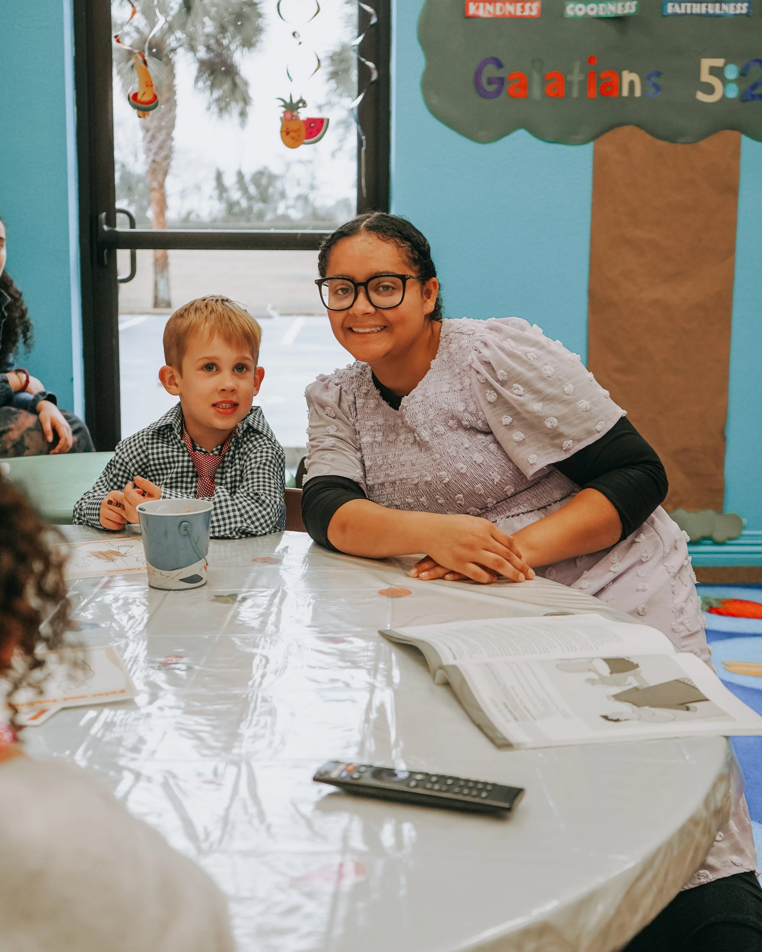 A woman and a young boy sitting at a table during a classroom activity or celebration. The table is covered with a clear plastic tablecloth, with papers and a remote control visible. In the background, a bulletin board displays colorful letters and words, including the names of books of the Bible and concepts like kindness, goodness, and faithfulness. A window reveals trees and an outdoor scene.