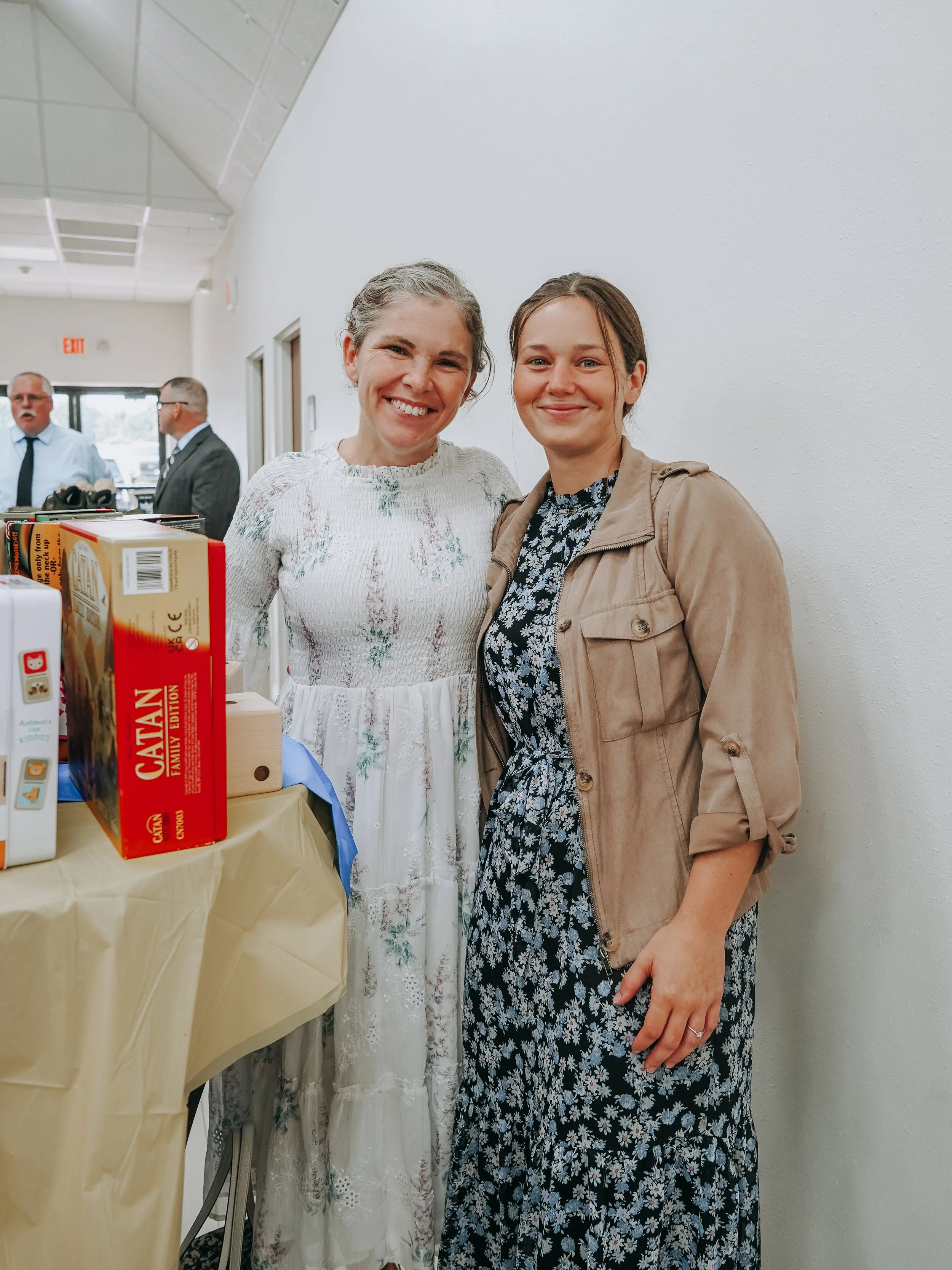 Two women smiling, standing next to each other in an indoor setting, with a table covered with a yellow cloth and some food boxes in front of them. There are other people in the background.