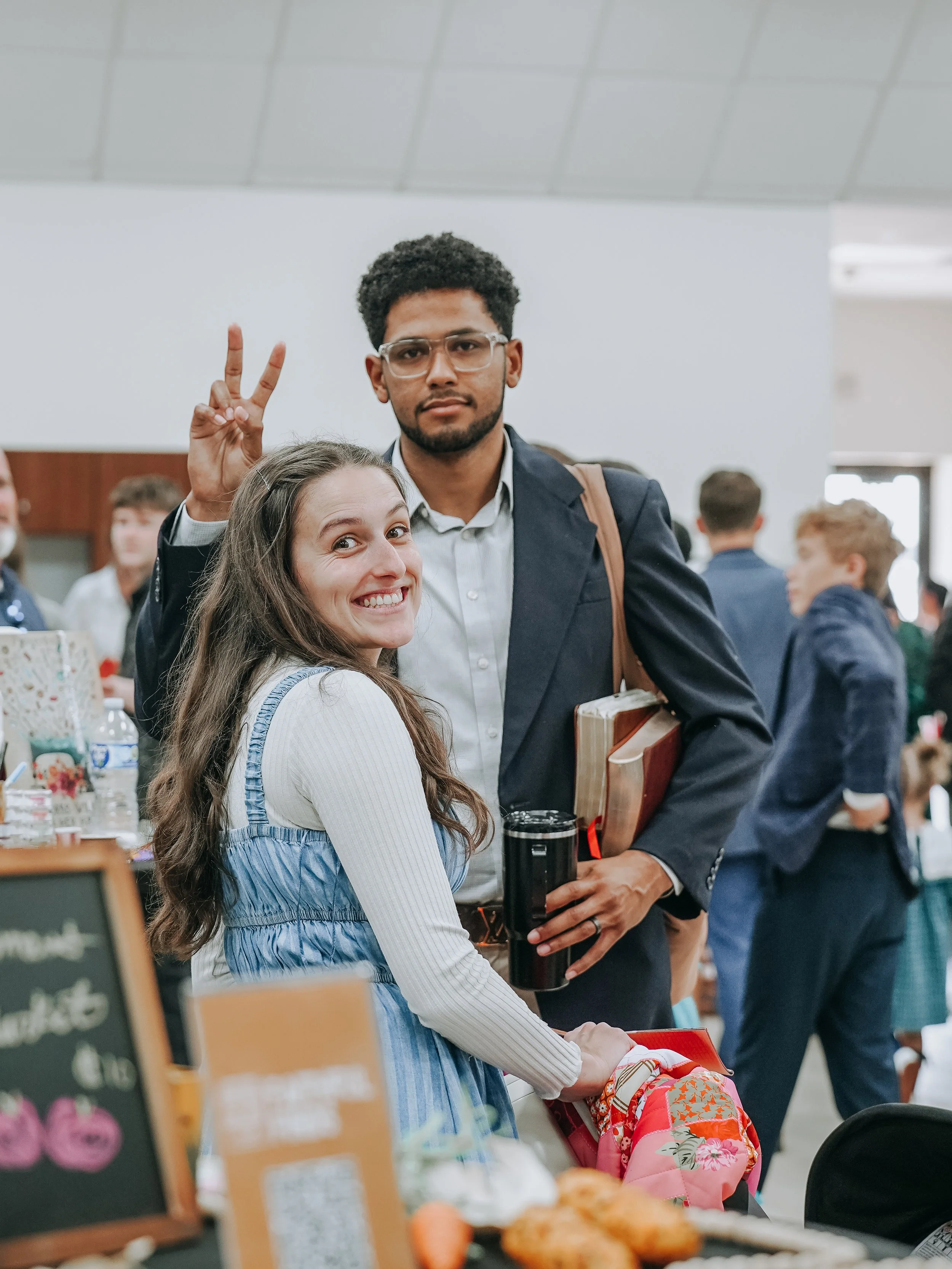 A woman with long brown hair and a man with glasses and curly hair, both smiling, in a crowded indoor setting. The woman is in the foreground, and the man is holding books and making a peace sign behind her. There are other people in the background, some interacting and some walking.