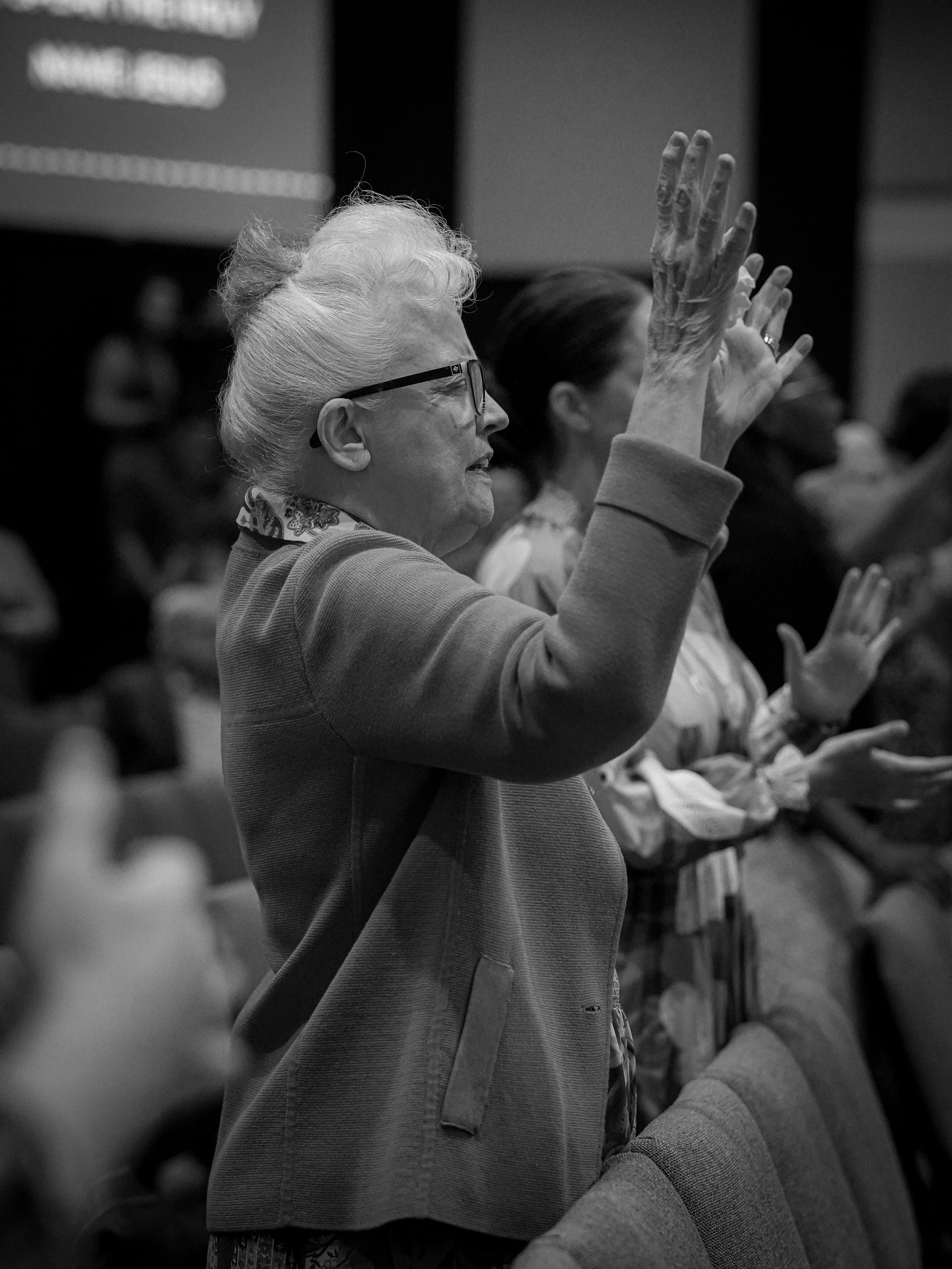 An elderly woman wearing glasses and a scarf, raising her hand with others around her in a group setting.