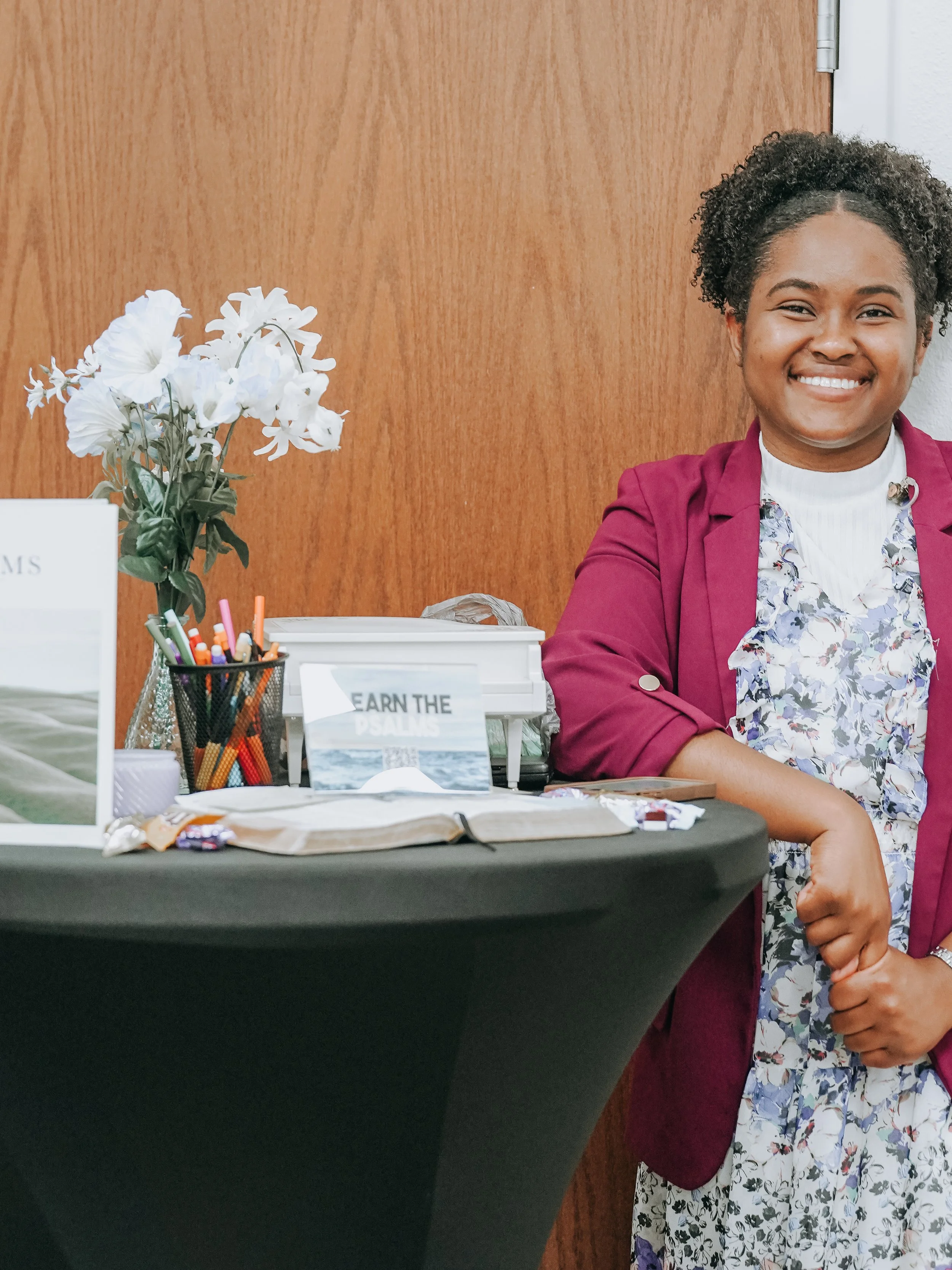 Smiling woman with curly hair wearing a maroon blazer and floral dress, sitting at a table with flowers and pamphlets, in front of a wood-paneled wall.