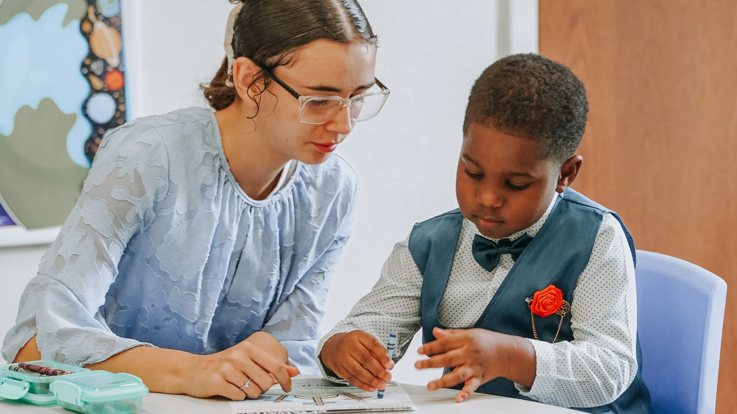 A woman with glasses and a young boy in a formal outfit sitting at a table, engaging in an activity together.