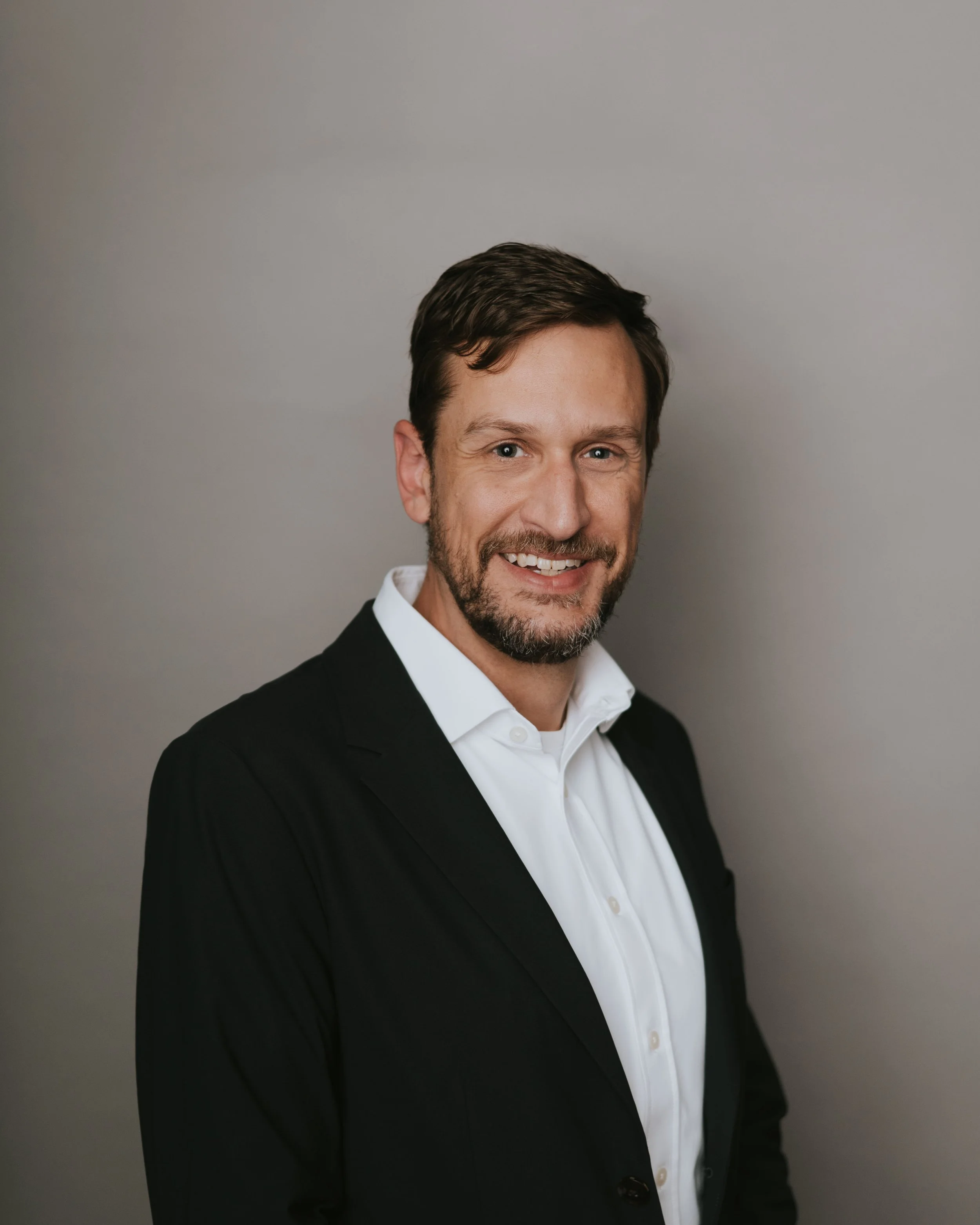 Headshot of a smiling man with brown hair, beard, dressed in a black suit and white shirt, against a plain gray background.