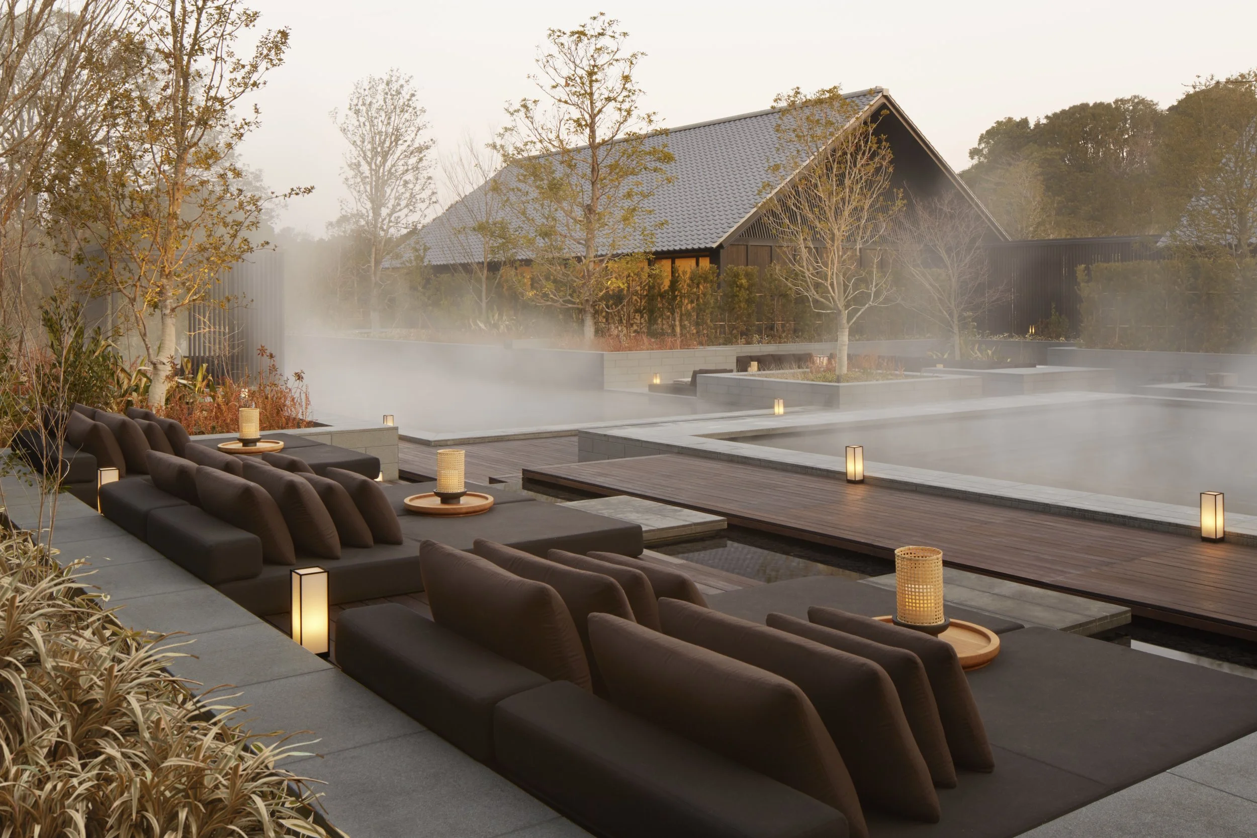 Modern outdoor hot spring with steaming water, surrounded by lounge chairs, small tables with candles, leafless trees, and autumn foliage fencing, with a house in the background.