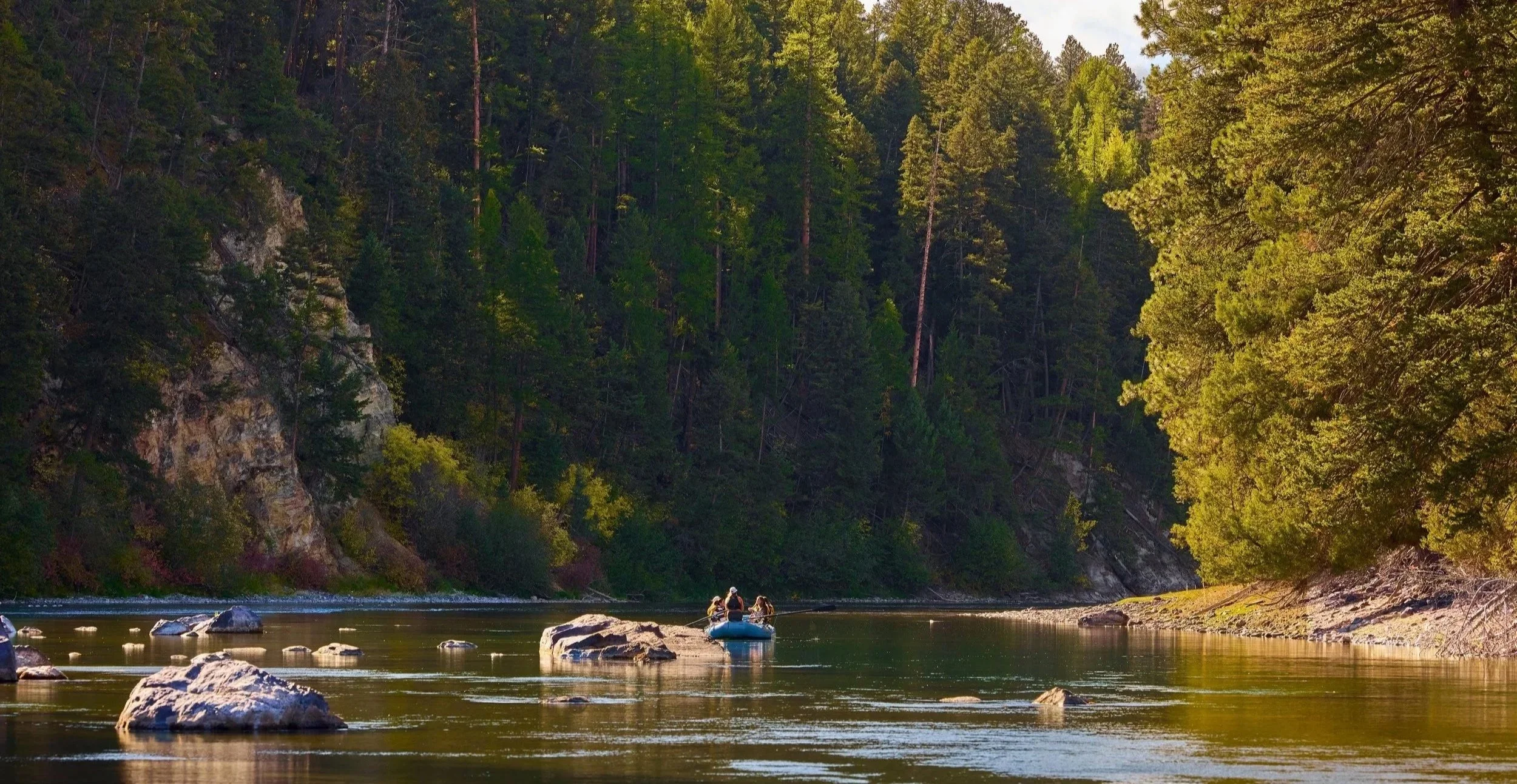 People kayaking on a calm river surrounded by dense green trees and rocky cliffs during daytime.