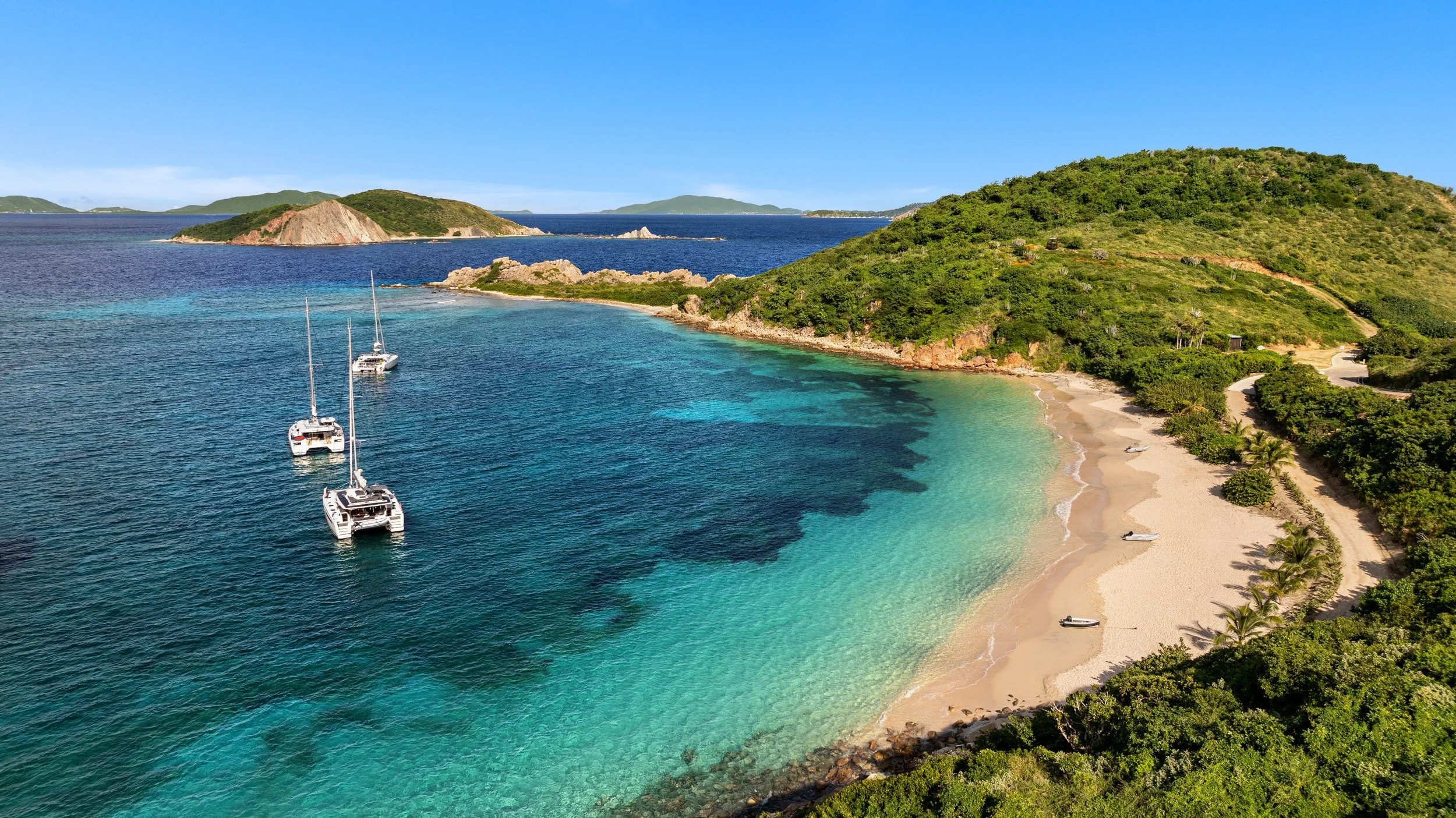 A scenic coastal landscape with green hills, sandy beach, turquoise water, and sailboats anchored in the bay.