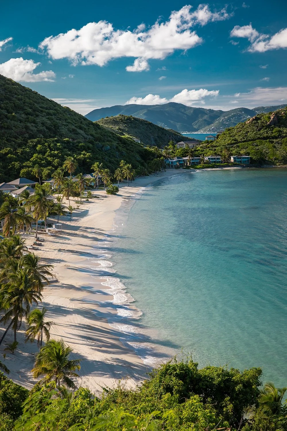 A scenic beach with white sand, palm trees, clear turquoise water, lush green hills, and mountains in the background under a partly cloudy sky.