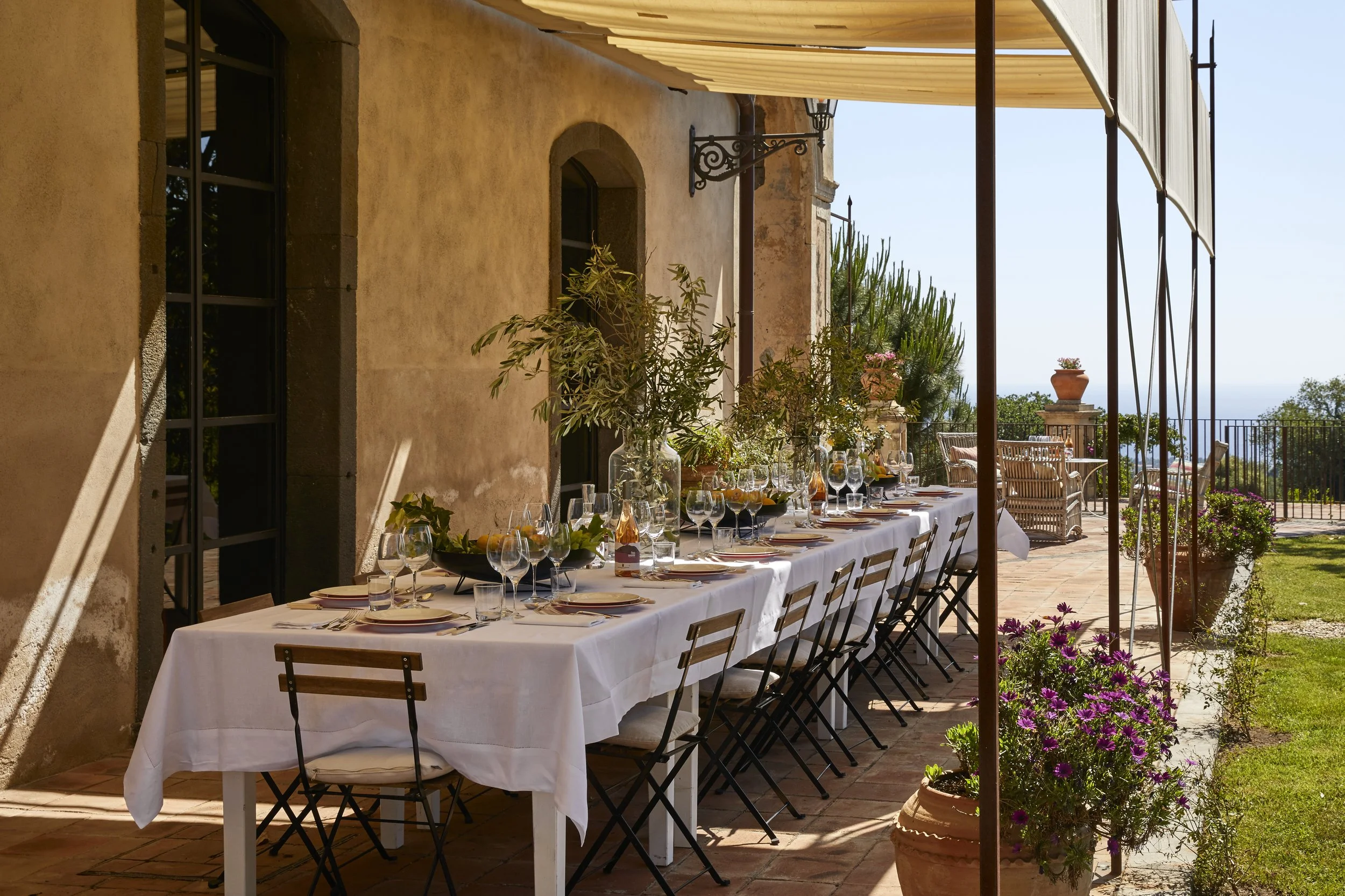 An outdoor dining setup with a long table covered with a white tablecloth, set with wine glasses, plates, and cutlery, under a partially shaded wooden pergola, with potted plants and a scenic view in the background.