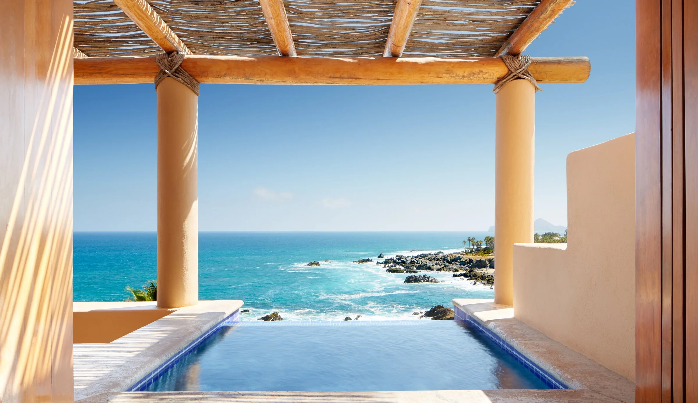 View through a veranda with wooden beams and columns overlooking the ocean, rocky shoreline, and distant mountains under a clear blue sky.