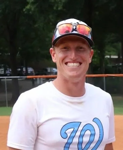 Smiling man wearing sunglasses on hat, white t-shirt with a blue logo, outdoors with a baseball field in the background.