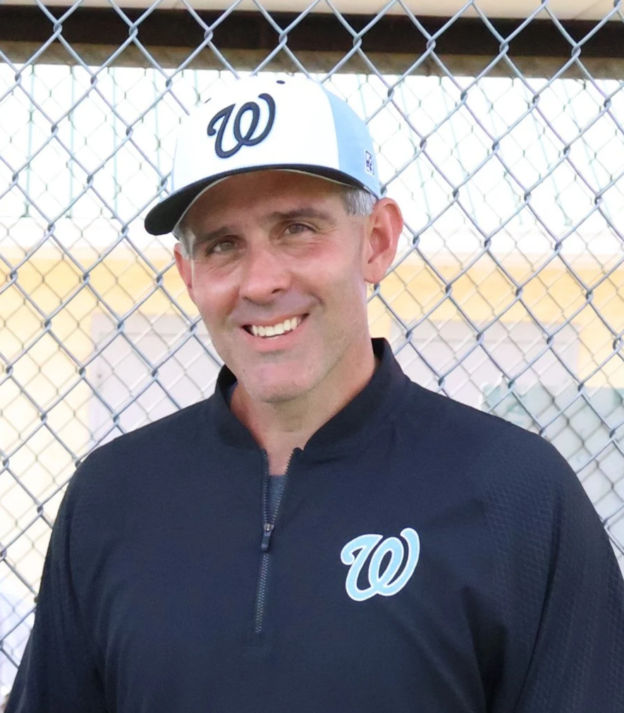 A man smiling wearing a West Walton All Stars baseball cap and dark sports jacket with a team logo, standing in front of a chain-link fence, likely on a baseball field.