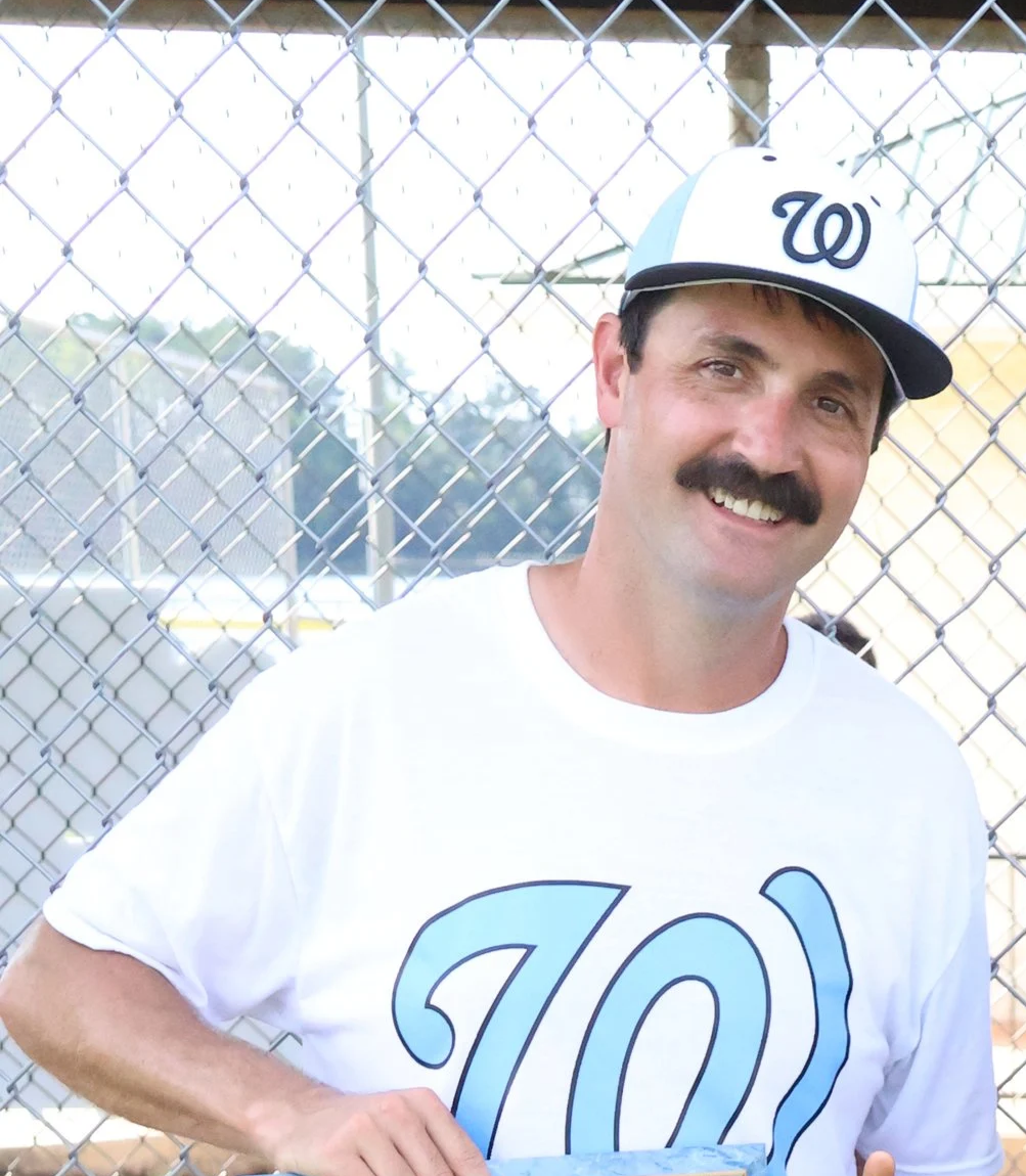 A man smiling at the camera wearing a West Walton All Stars baseball cap and a white T-shirt with a large blue 'W' logo. He is standing in front of a chain-link fence.