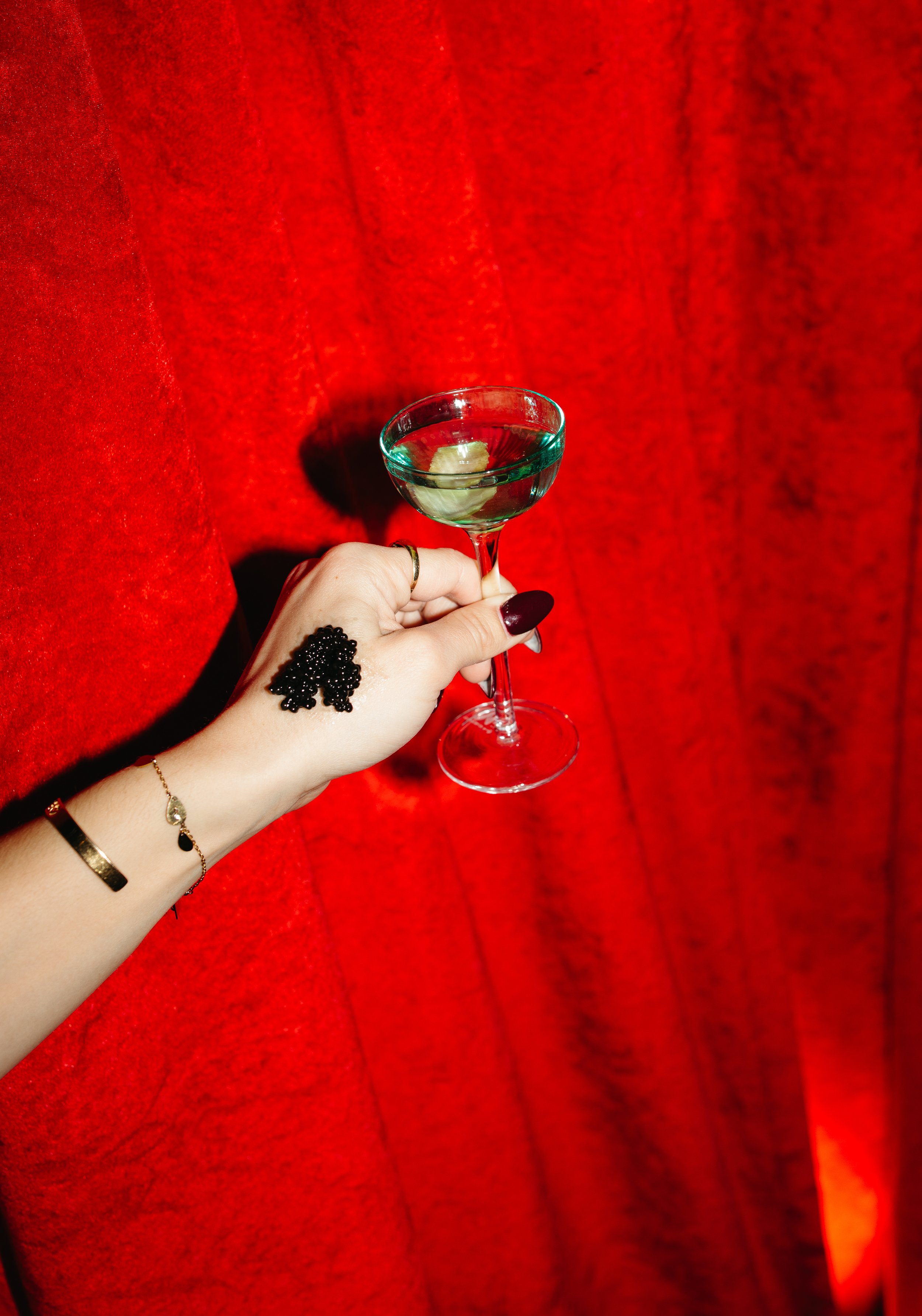 A hand holding a cocktail glass with a clear liquid and a lime slice against a red velvet background.
