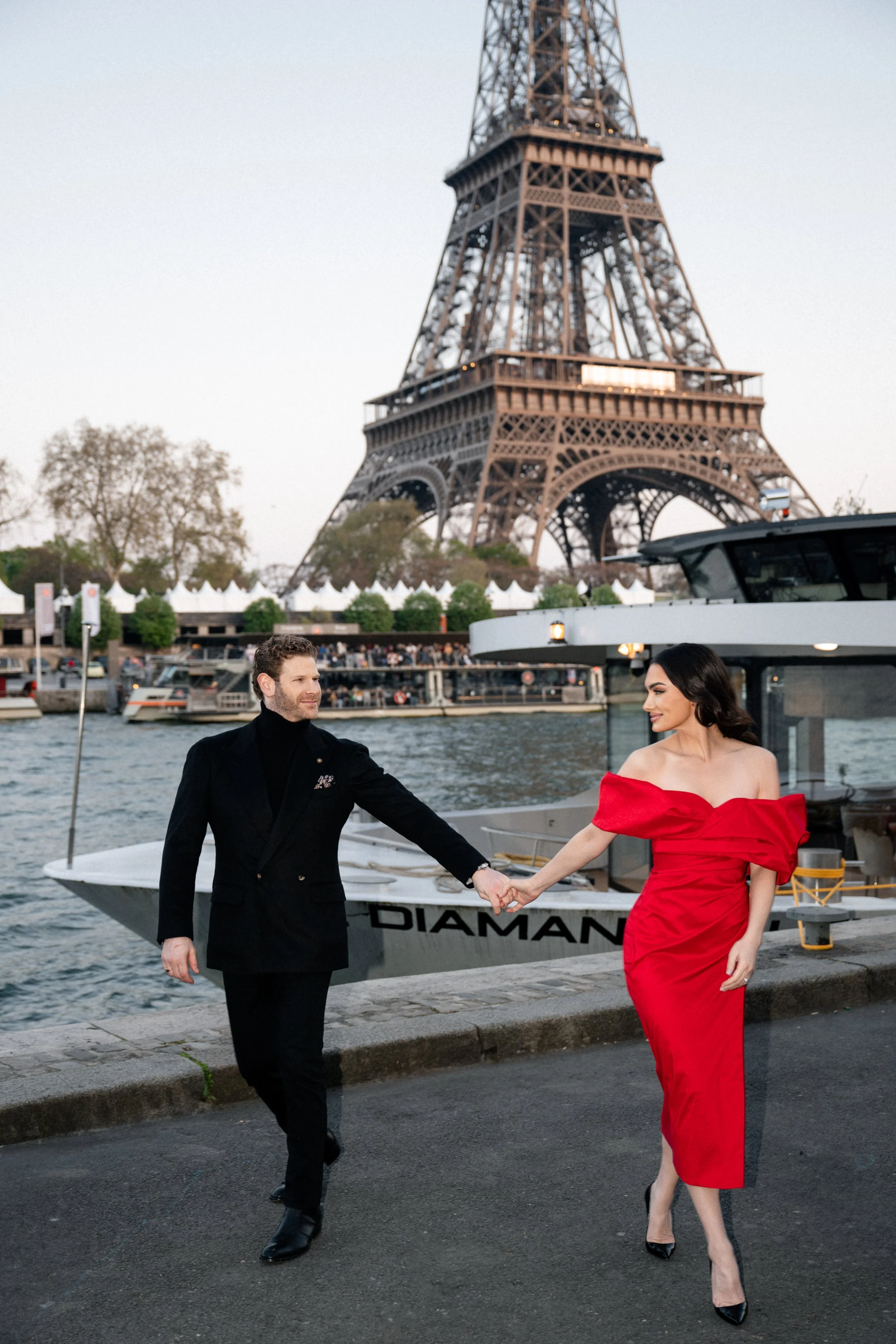 A man and woman holding hands on a dock in front of the Eiffel Tower with boats on the river.