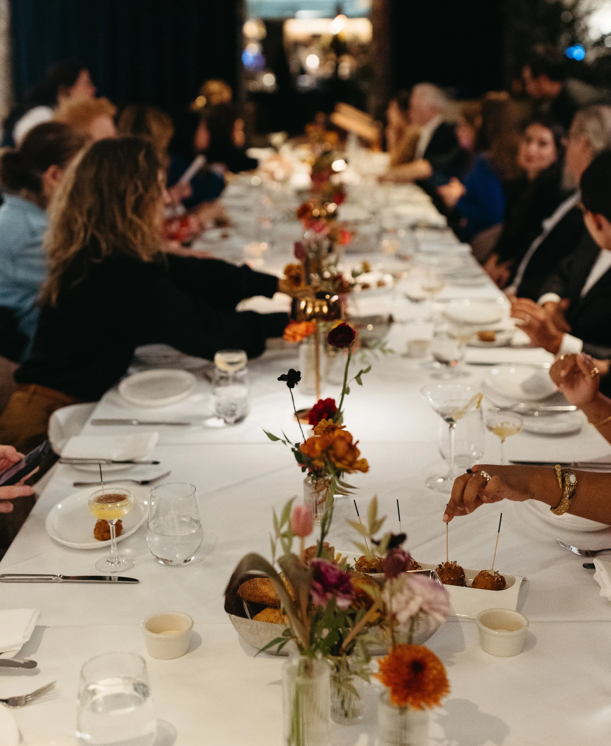 A group of people sitting around a long dining table at a formal gathering or dinner event, with floral centerpieces and glassware.