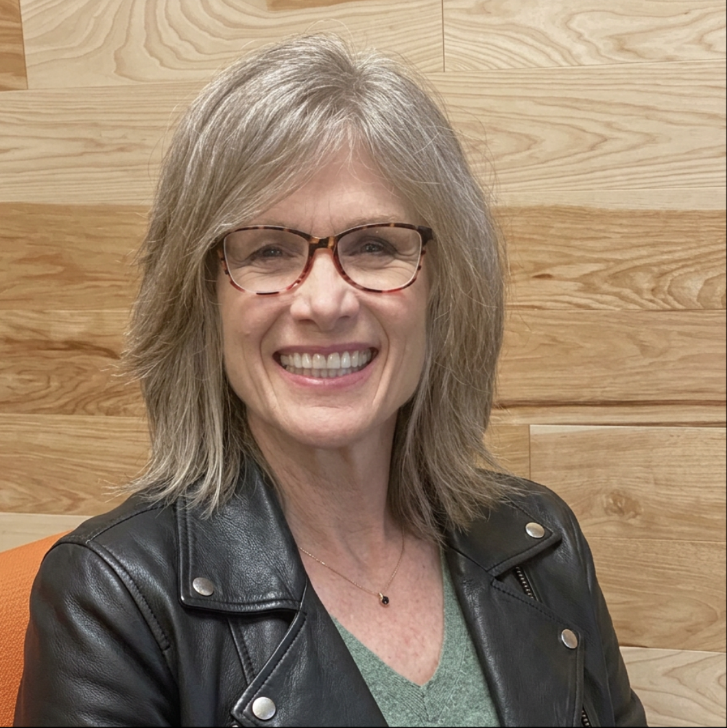 A smiling middle-aged woman with shoulder-length gray hair, wearing glasses, a black leather jacket, and a green top, sitting on an orange chair against a wooden wall background.