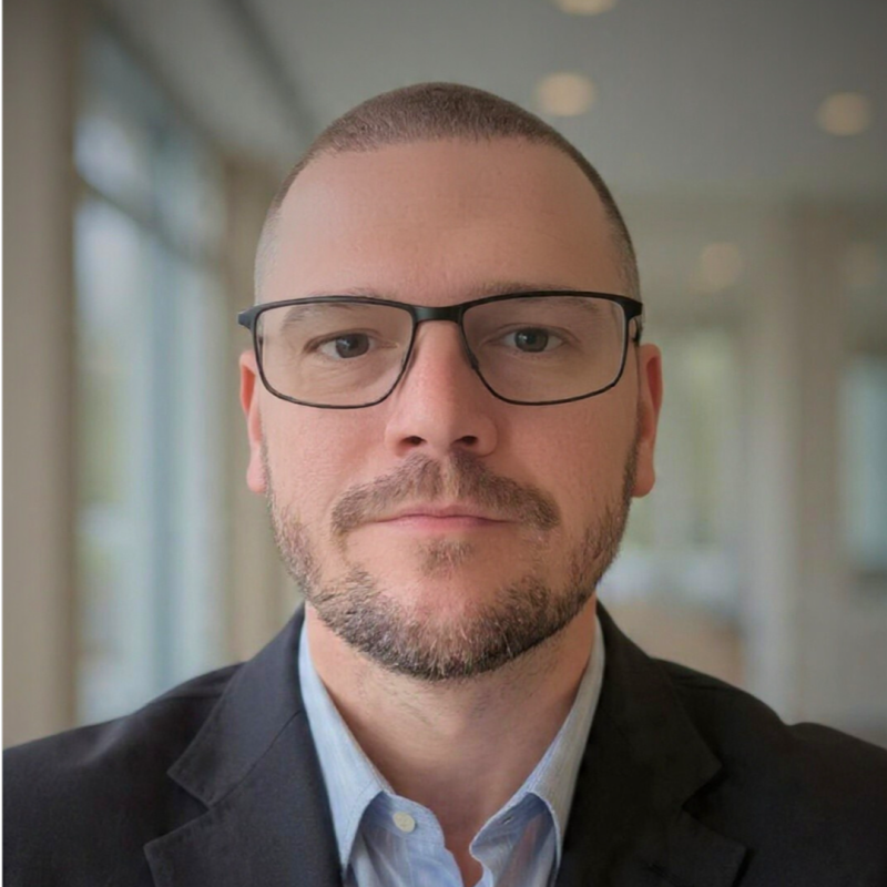 A photo of William Stetson, founder and chief executive officer of Dad Can Help Handyman Services wearing glasses, short hair, in a dark suit and light blue shirt, standing indoors with blurred windows in the background.