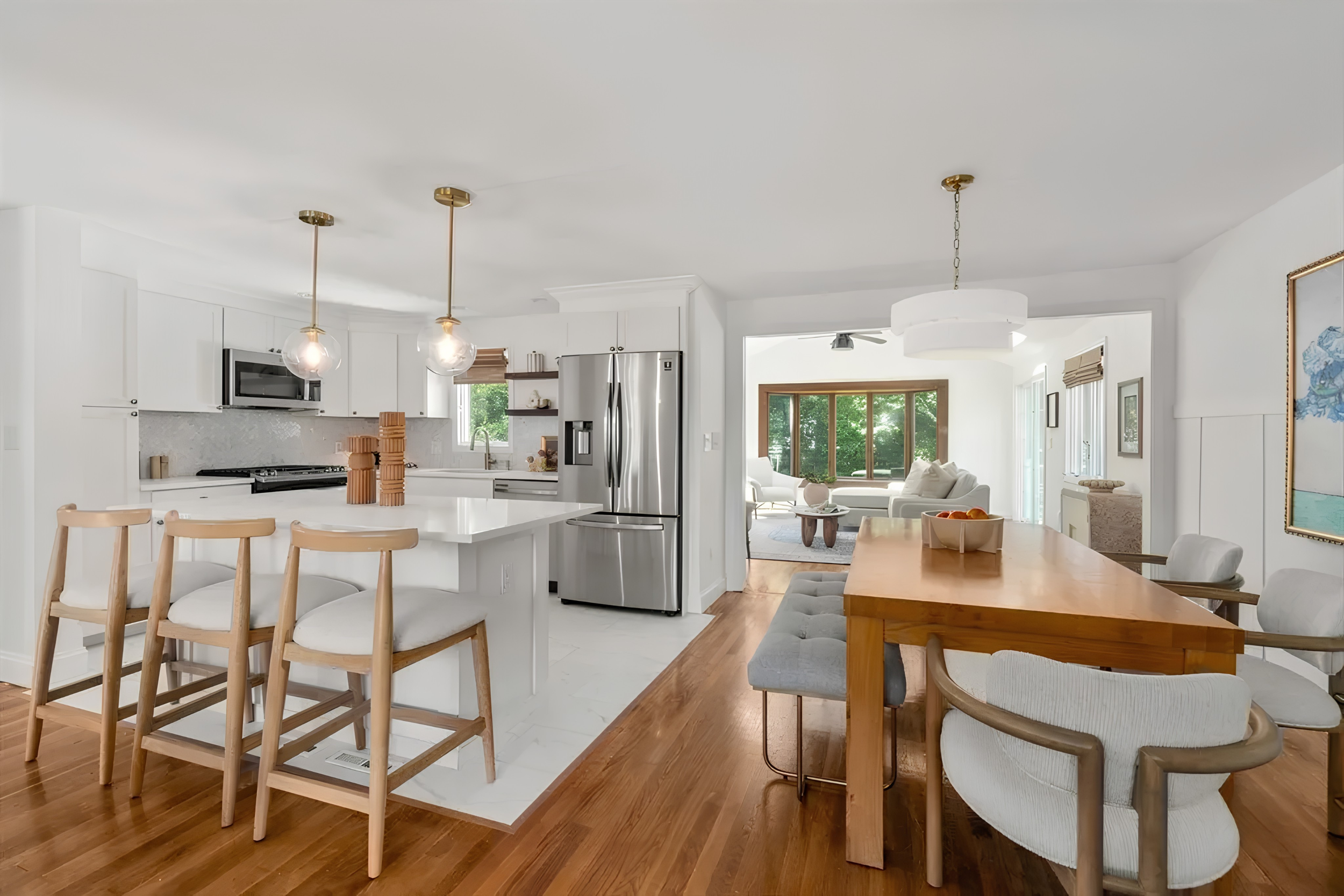 Open-concept kitchen and living room with white cabinets, stainless steel appliances, wooden dining table, beige chairs, and large windows showing green trees outside.