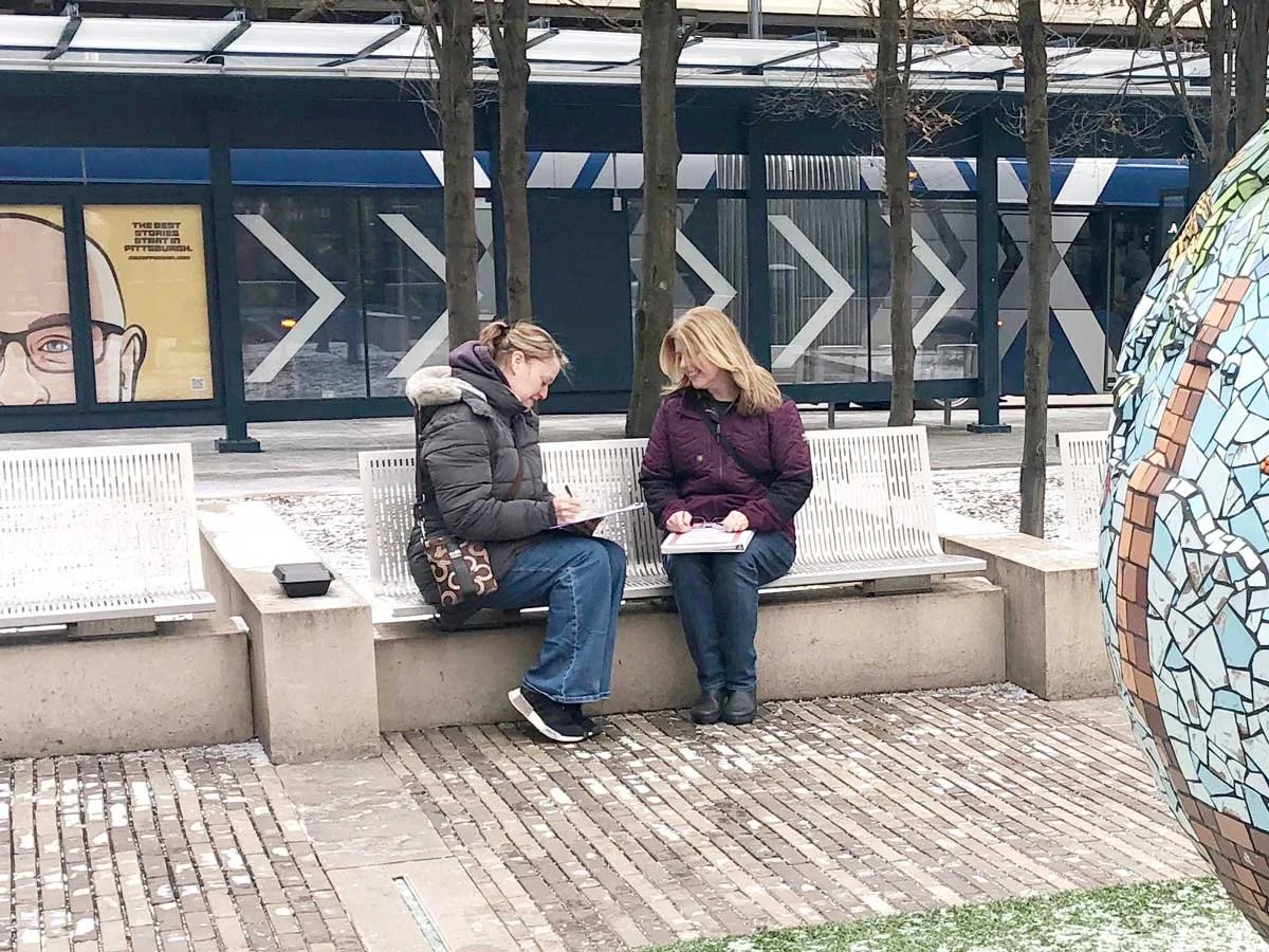 Artist Marsha Rollinger sitting on a bench outside in conversation with a woman from one of the participating PittsBirds communities.