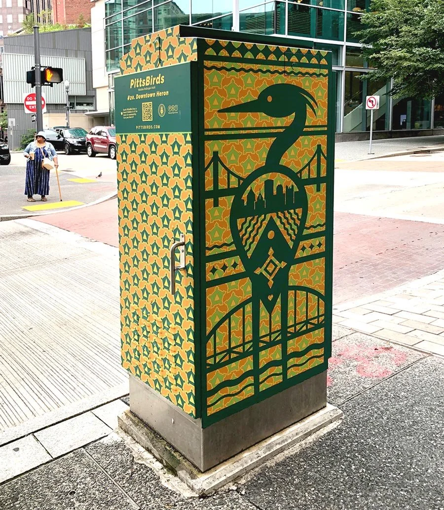 Decorative electrical box in an urban area featuring a design of a bird, a bridge, city skyline, and landscape in black outline on a yellow and green patterned background.