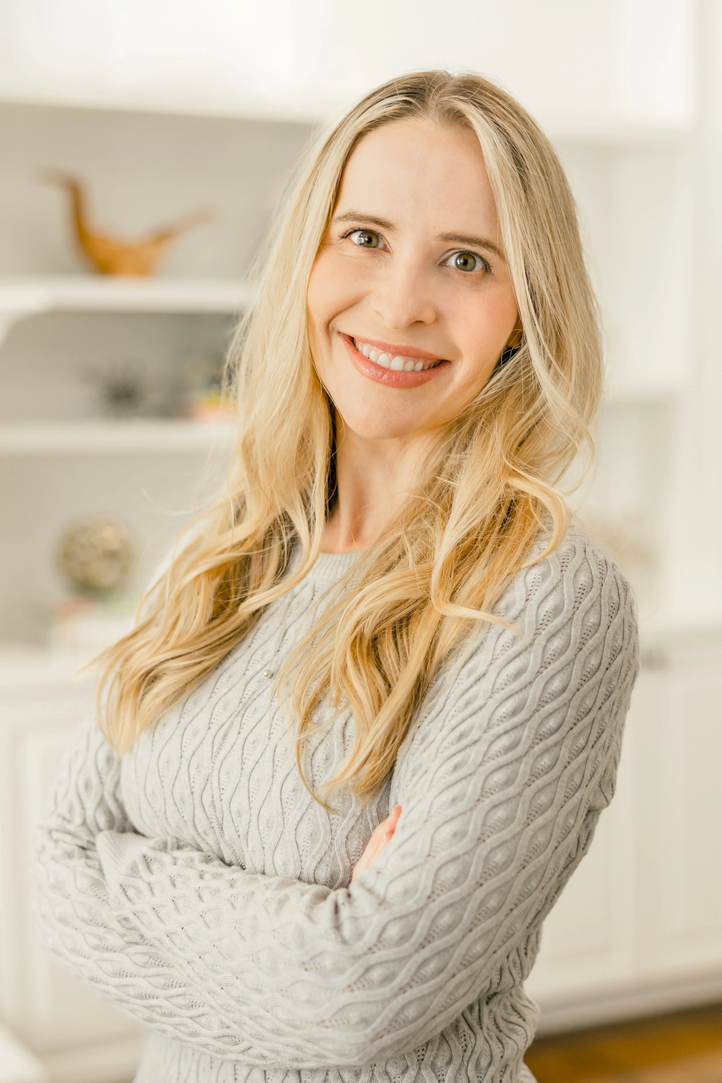A young woman with long blonde hair, smiling, standing in a well-lit room with blurred background, wearing a light-colored cable-knit sweater.