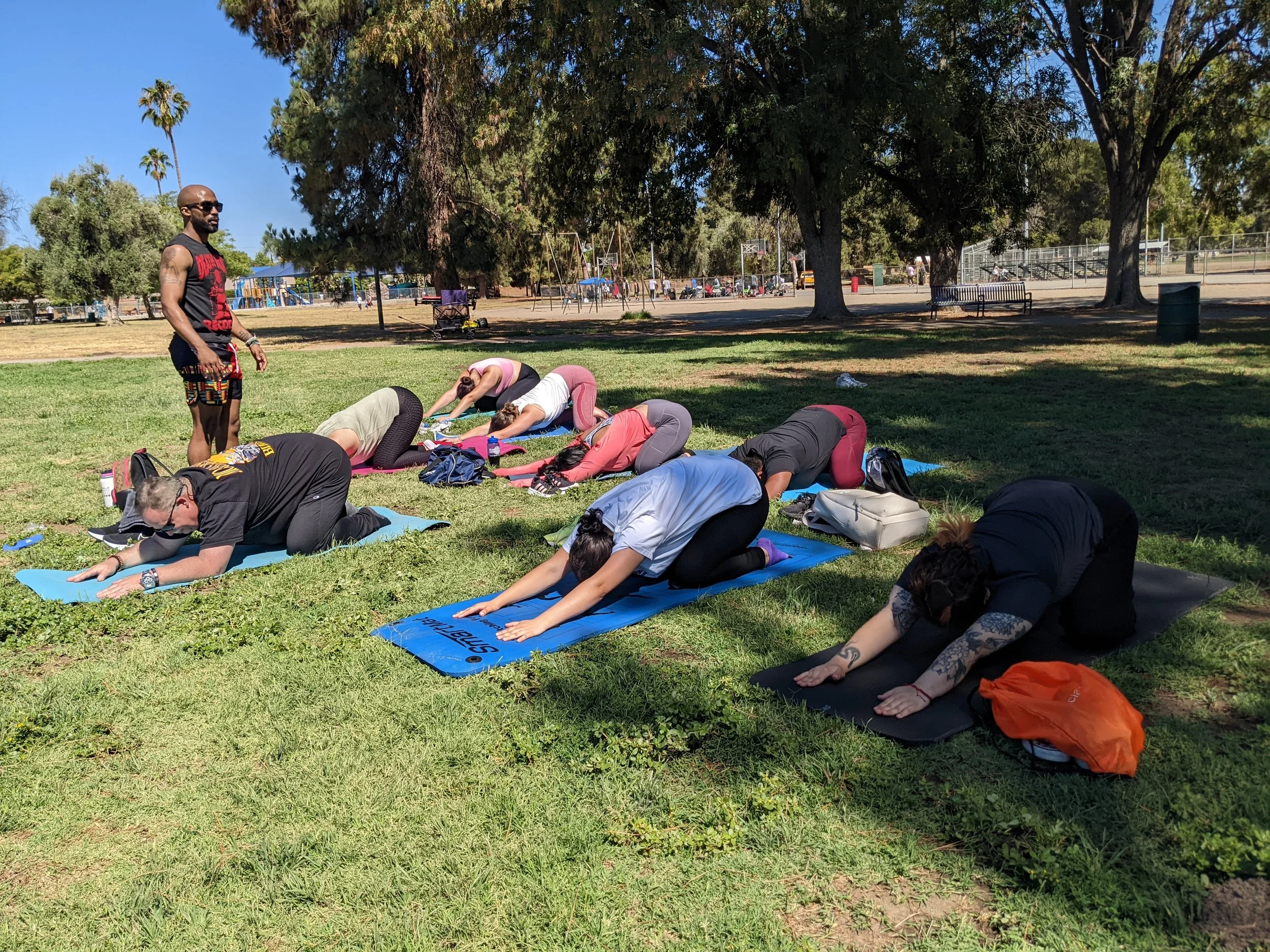 People practicing yoga outdoors on yoga mats in a park with trees, a playground, and a basketball court in the background on a sunny day.