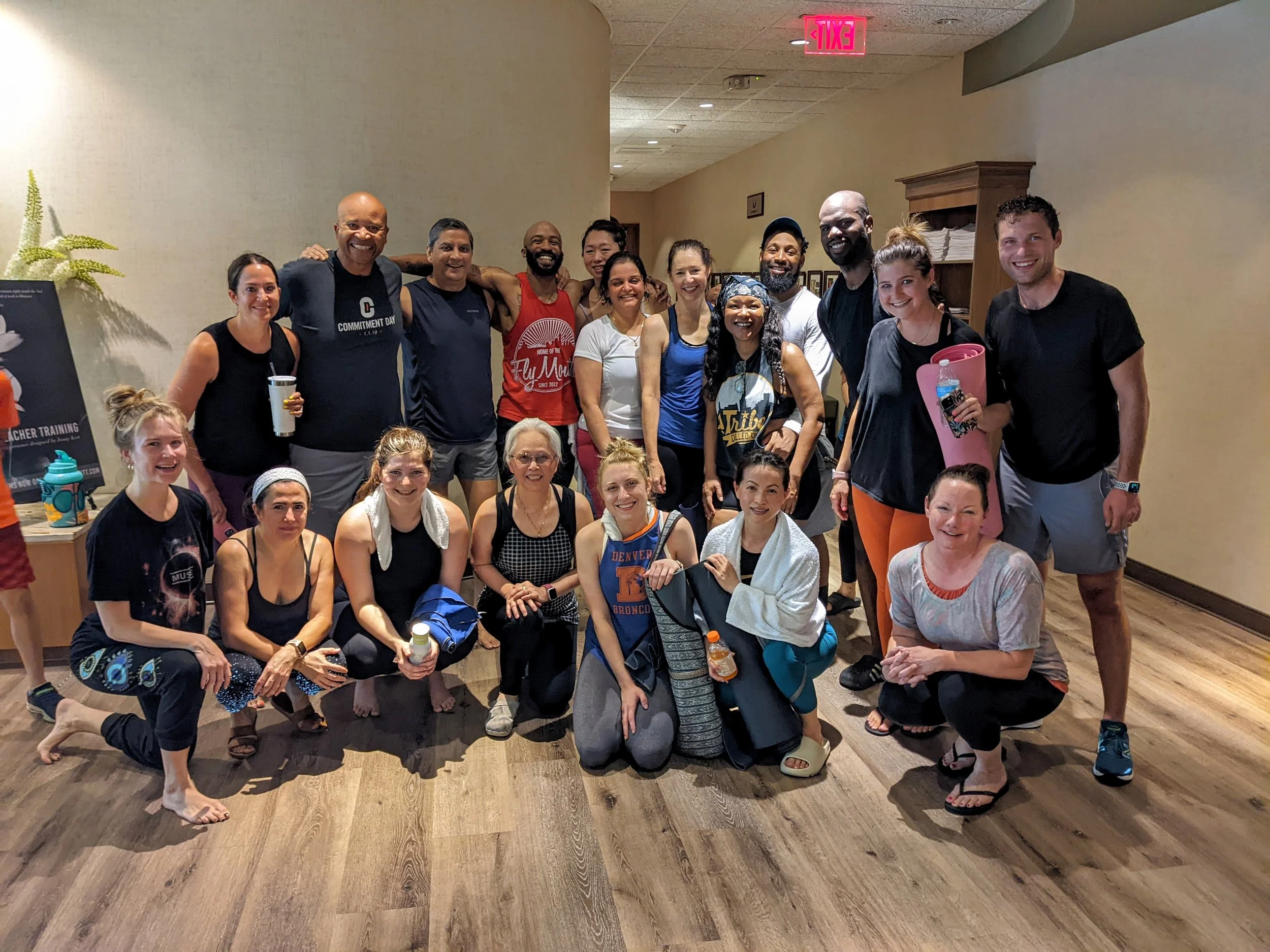 A group of people gathered indoors, some standing and some sitting, smiling at the camera. The group is diverse, with men and women of various ages and ethnicities. The room has wood flooring and beige walls, with an emergency exit sign visible on the ceiling.