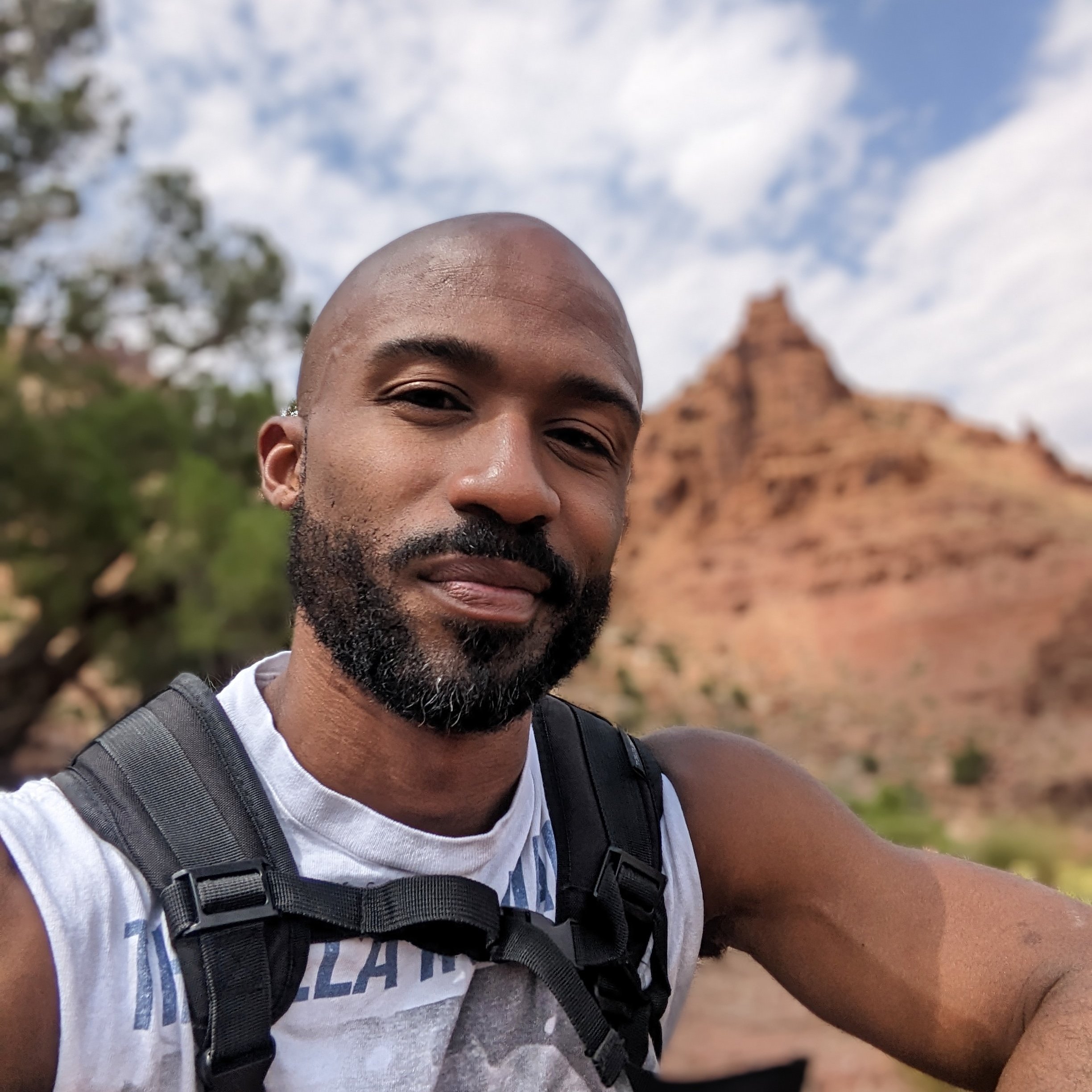 A close-up of a man's face outdoors, smiling, with a bald head and beard, wearing a backpack and a white T-shirt with blue lettering, against a background of trees, blue sky, and a red brick structure.