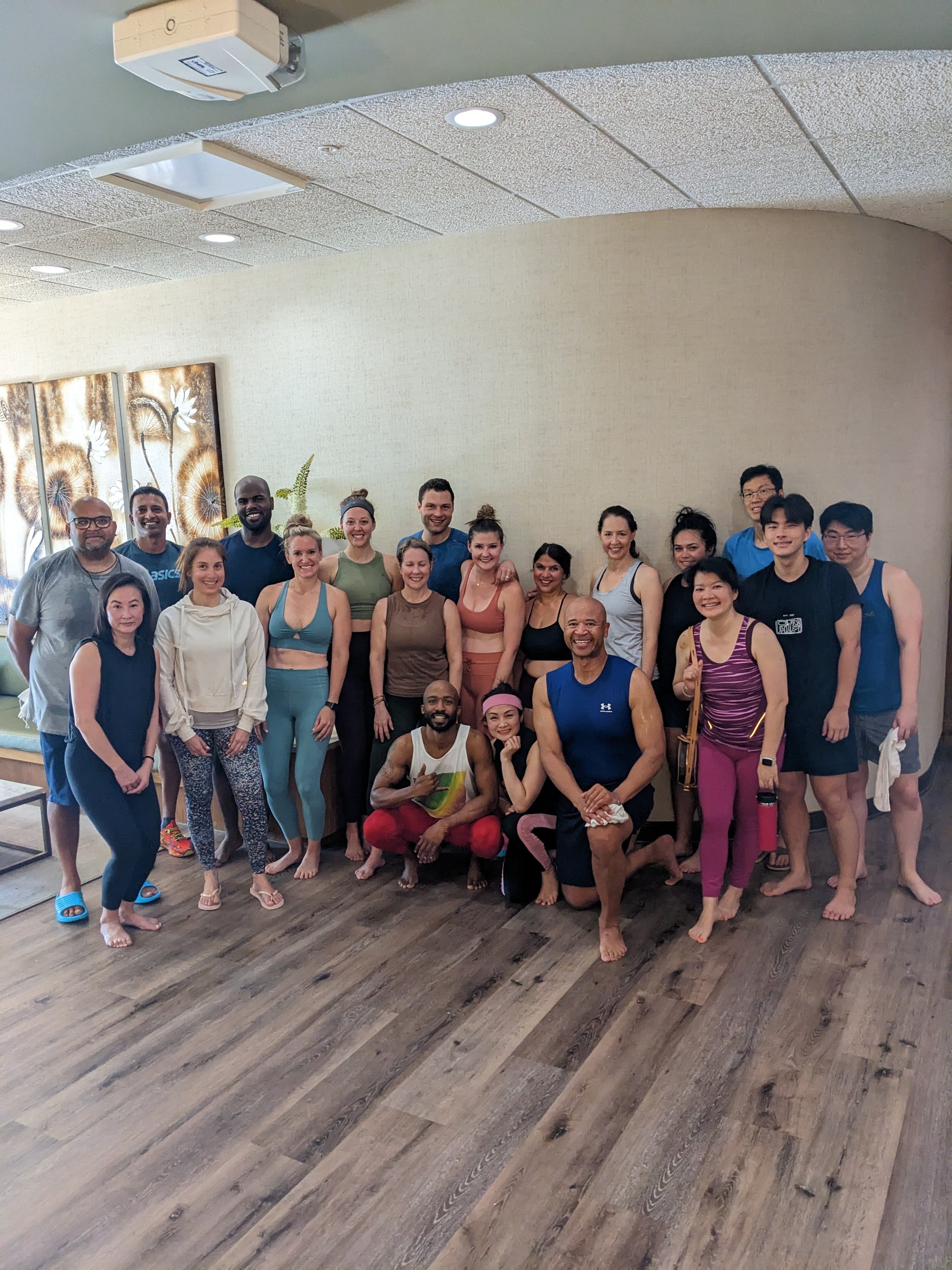 A diverse group of people in workout attire, smiling and posing together in a fitness or yoga class room.