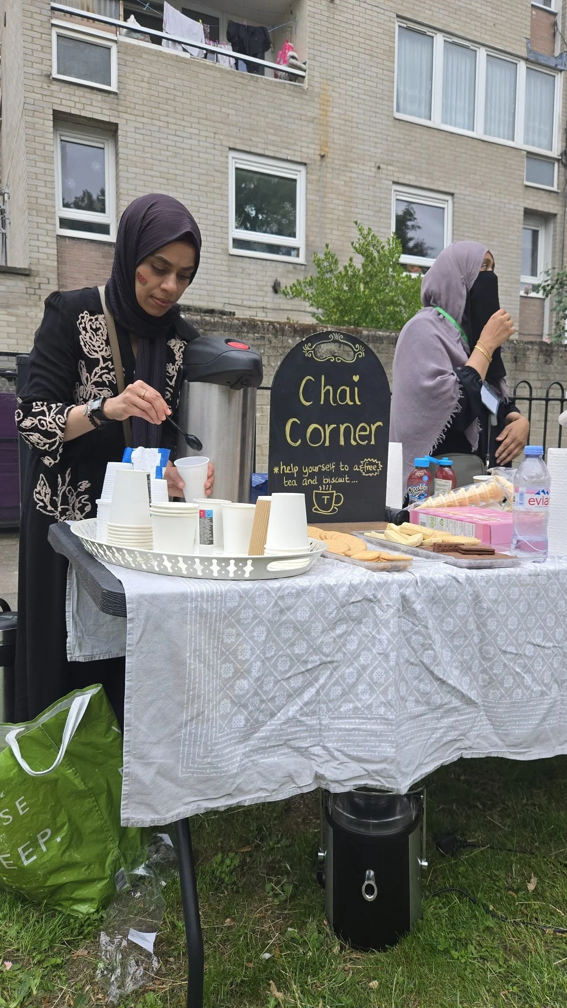 Two women at a small outdoor tea and biscuit stand called 'Chai Corner.' One woman is pouring tea from a large thermos, and the other is sipping tea. The table has disposable cups, water bottles, biscuits, and packaged snacks. There is a blackboard sign with yellow writing and a teacup drawing, advertising free tea and biscuits. The stand is set up on grass in front of an apartment building.