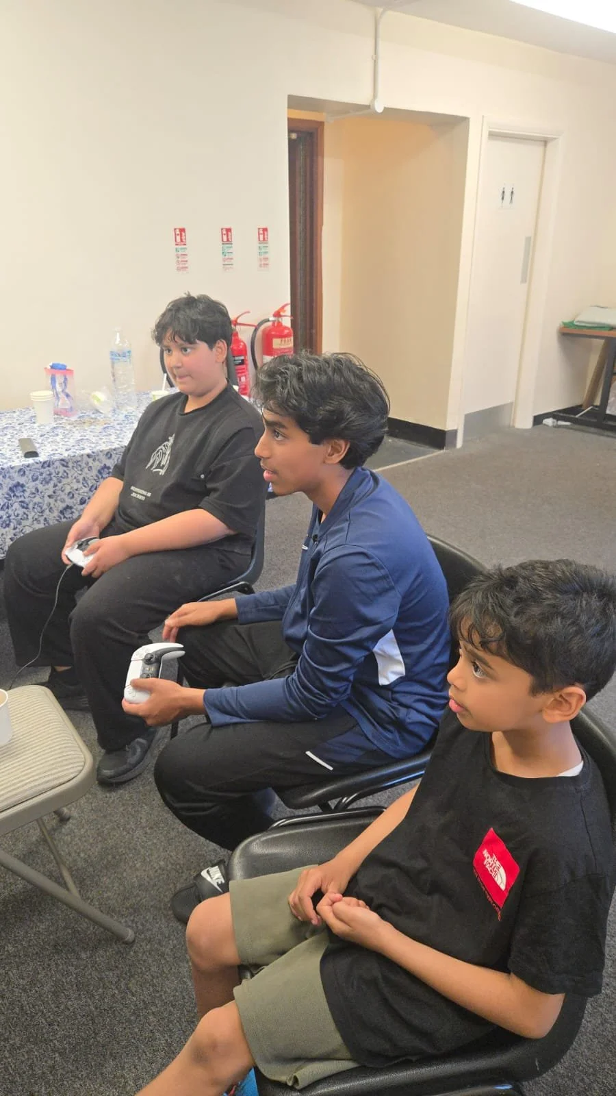 Three young boys sitting in chairs watching a screen, with two holding gaming controllers.