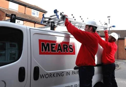 Two workers in red uniforms and white helmets installing equipment on a company van with MEARS logo