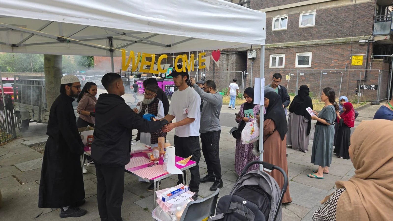 People standing in line at an outdoor food stall under a white canopy with a yellow 'WELCOME' sign, in an urban area with brick buildings in the background.