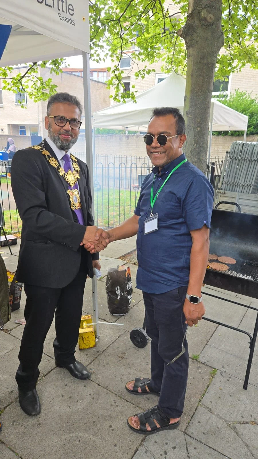 Two men shaking hands at an outdoor event, standing near a barbecue grill and trees.