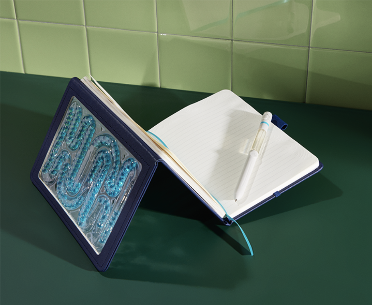 Open notebook with lined pages, white ballpoint pen with a blue clip, and a blue folder with alphabet beads spelling 'NOW' on a green surface with green tiled wall in background.