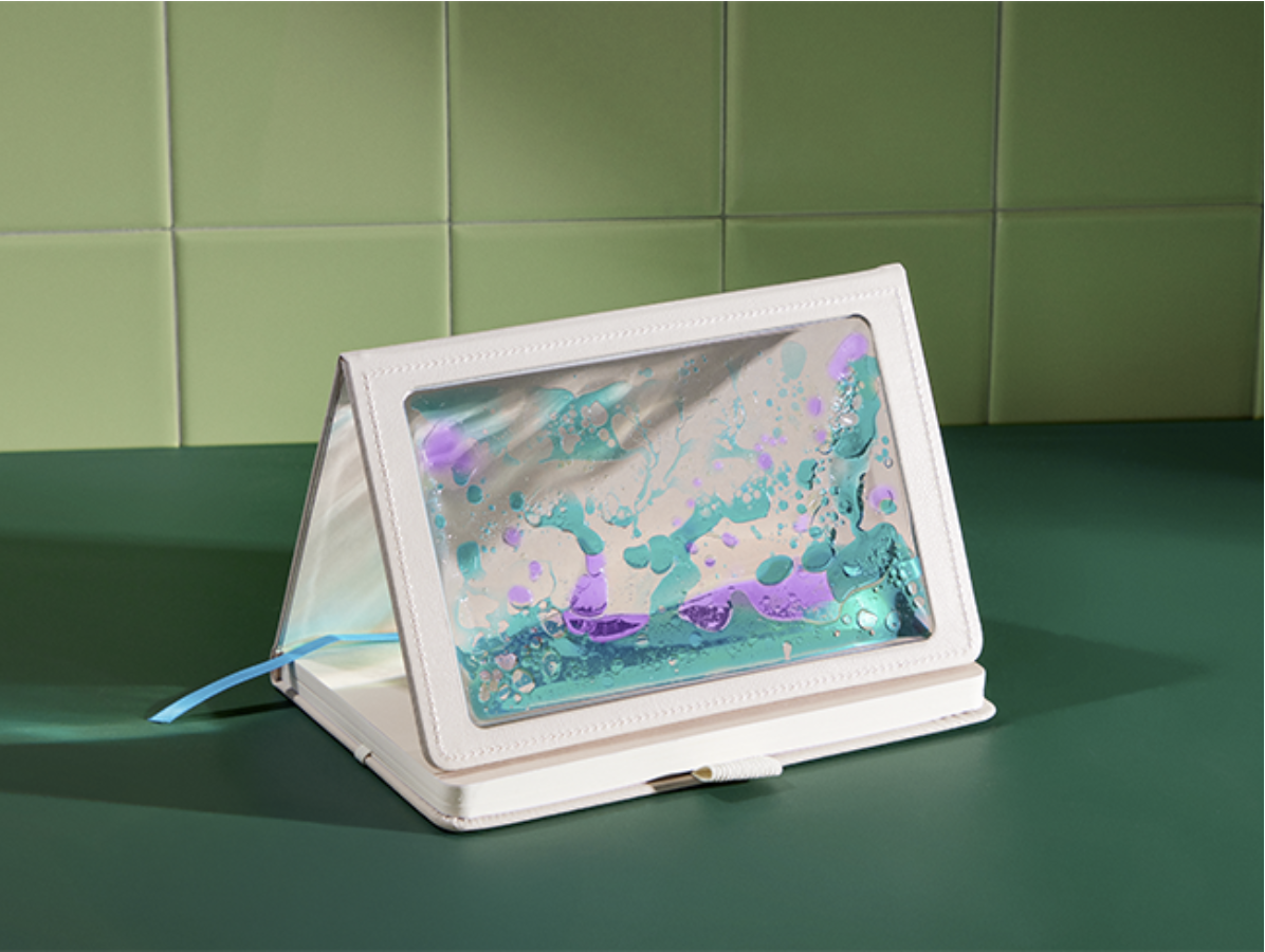 A foldable white notebook or diary with a transparent cover displaying colorful ink splashes in purple, blue, and teal, sitting on a green surface against a green tiled wall.