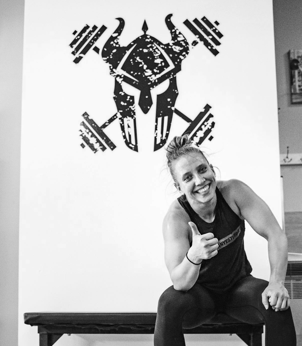 A woman with a ponytail, smiling and giving a thumbs-up, sitting on a bench in front of a wall with a large black graphic of a stylized animal skull with horns and abstract shapes.