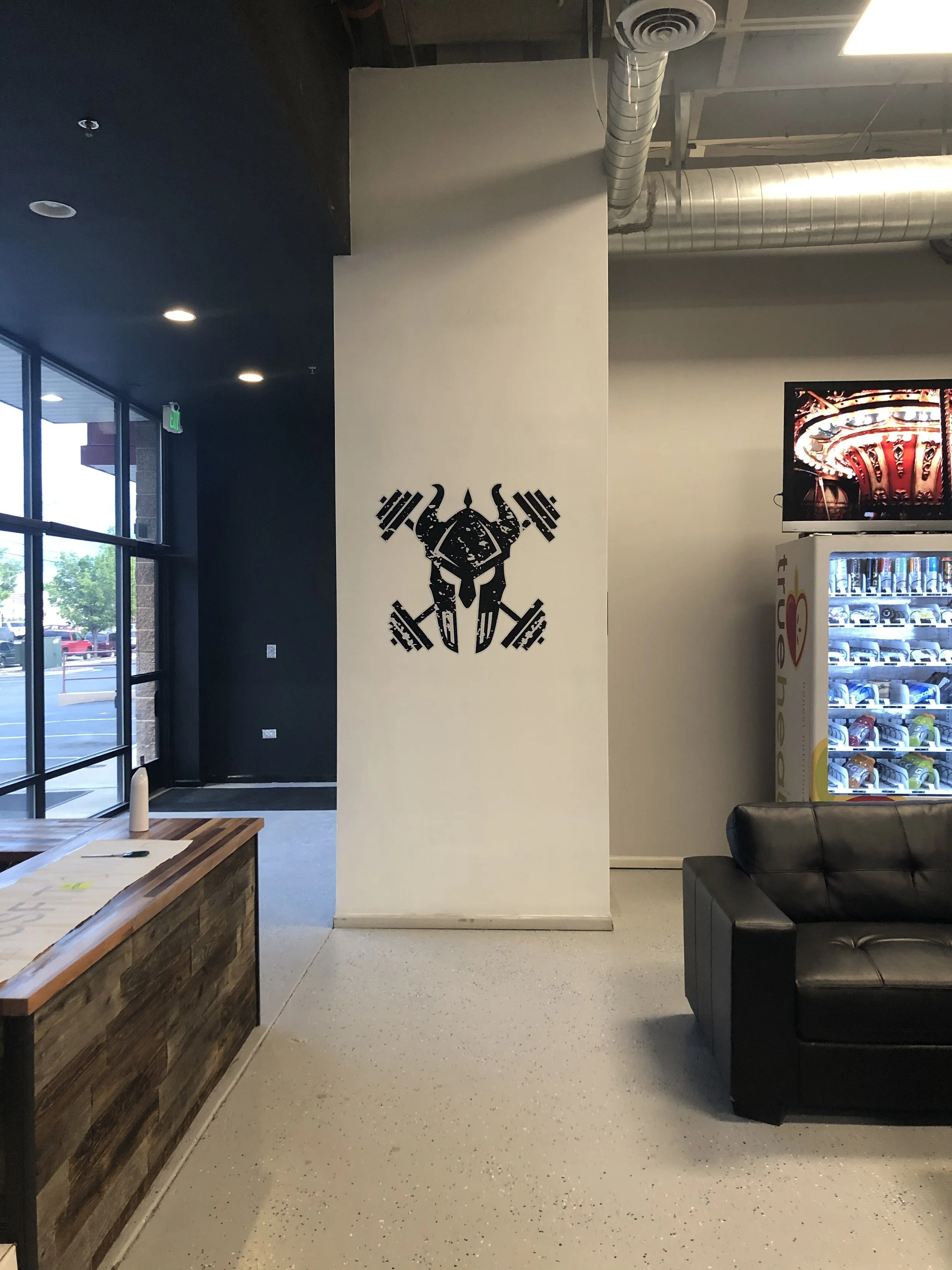 Interior of a modern building with a dark ceiling, illuminated ceiling lights, large windows, a wooden reception desk, a white wall with a black graphic of a robotics or mechanical design, a vending machine with drinks, a black leather couch, and duc