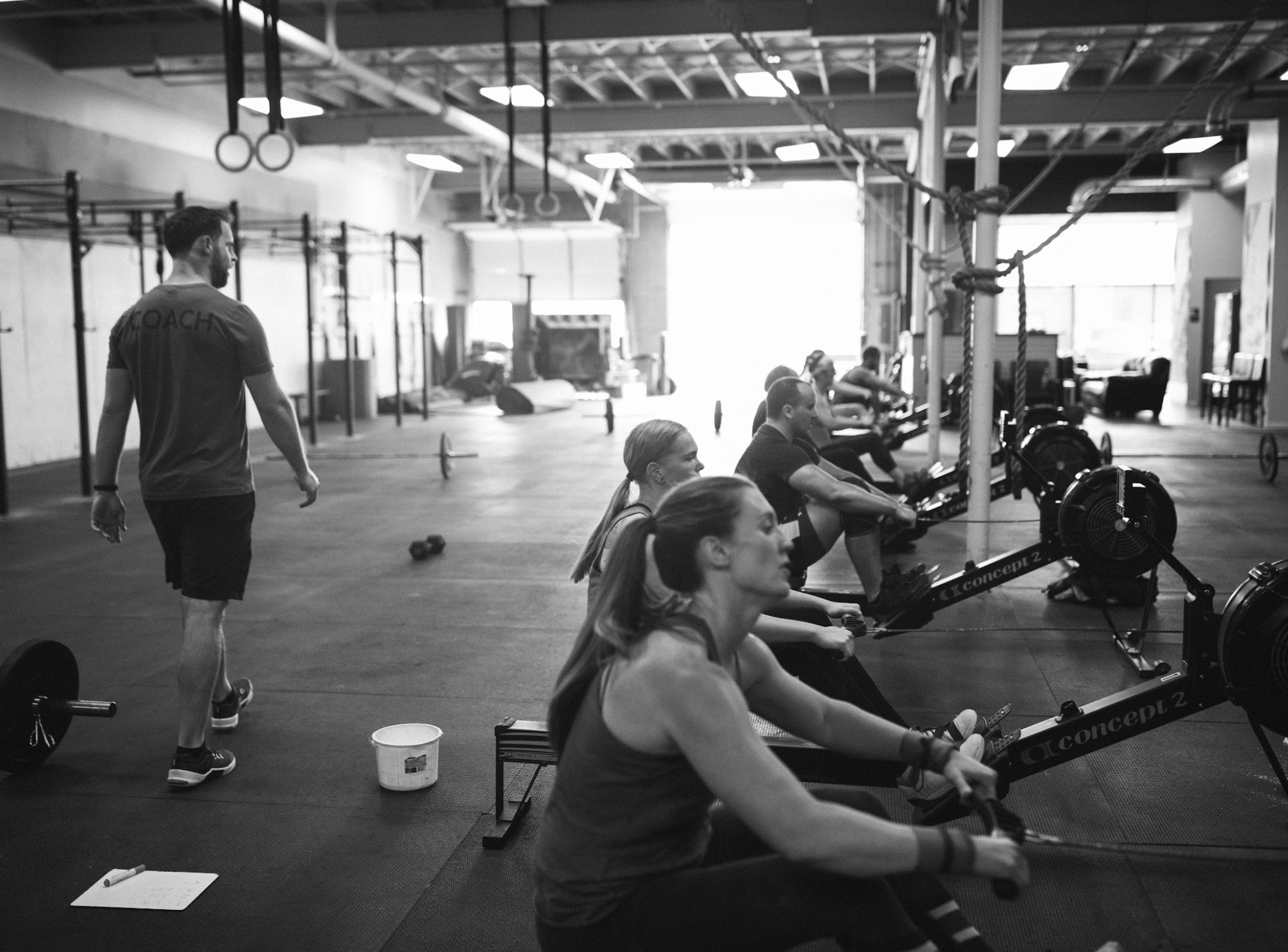 A group of people participating in a rowing exercise on indoor rowing machines in a gym. An instructor stands nearby observing.