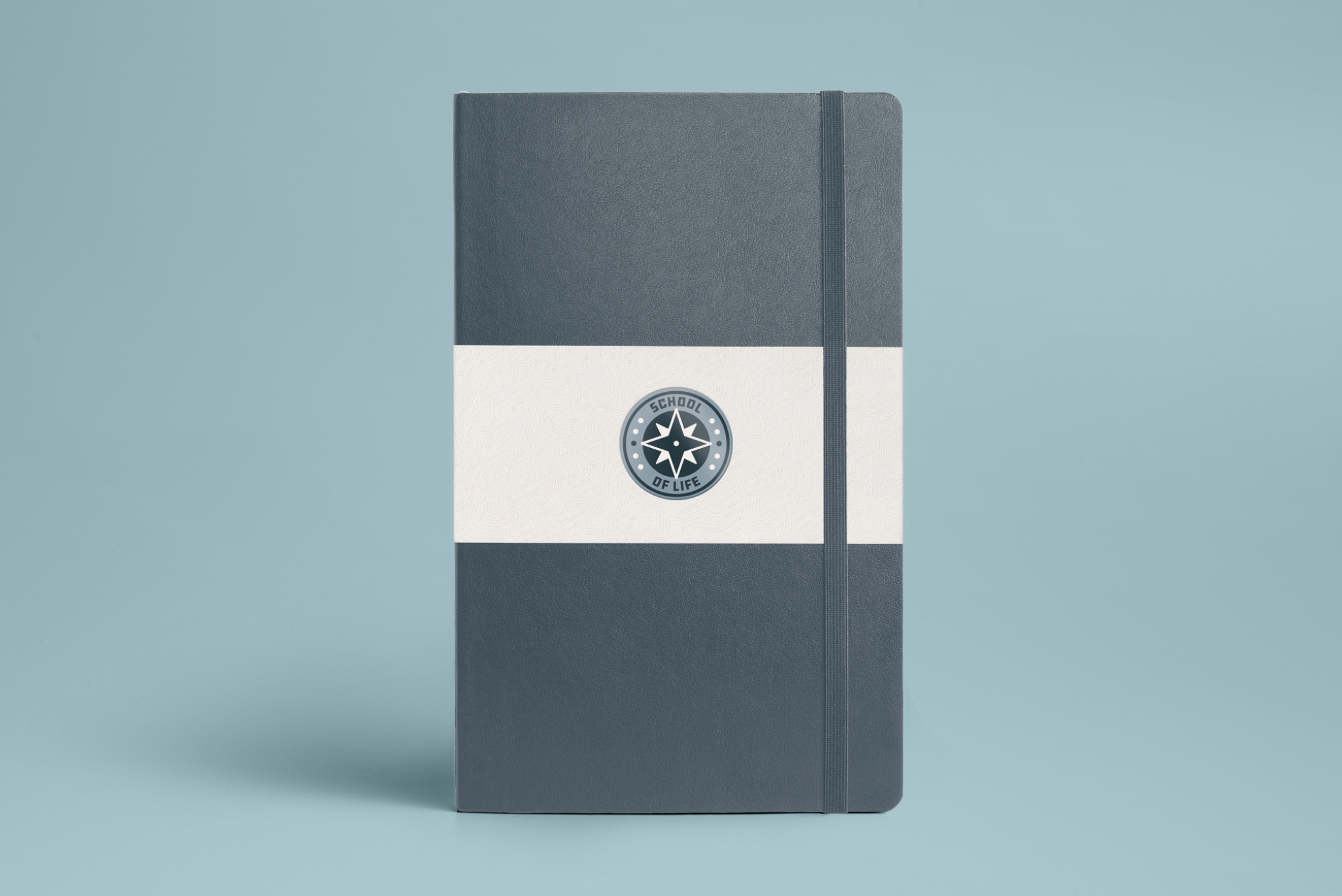 A gray hardcover notebook with a black elastic band, placed against a light blue background. The lower part of the cover features a white band with a circular logo reading 'School of Life' with a star symbol.