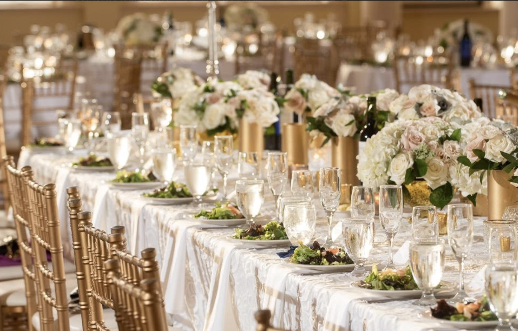 Elegant banquet table with white floral centerpieces, wine bottles, glasses, and place settings in a decorated event hall.