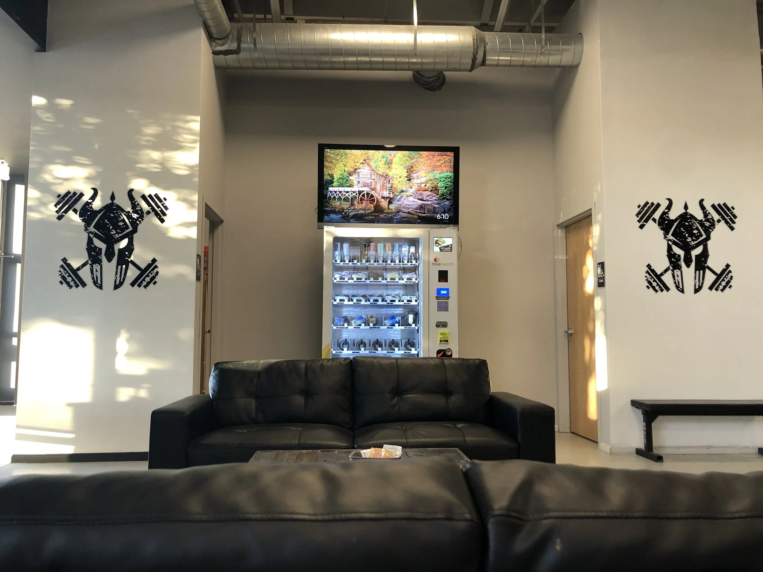 A waiting area or lobby with a black leather couch in the foreground. Behind it, a vending machine with snacks and drinks. On the wall above, a flat screen monitor displaying a scenic image of a watermill in a forest.