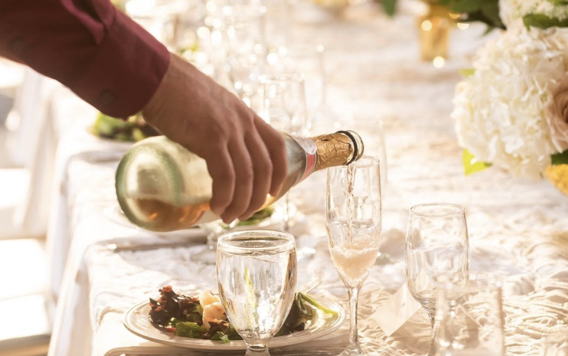 A person pouring champagne into a glass at a celebration or event table decorated with flowers and glassware.