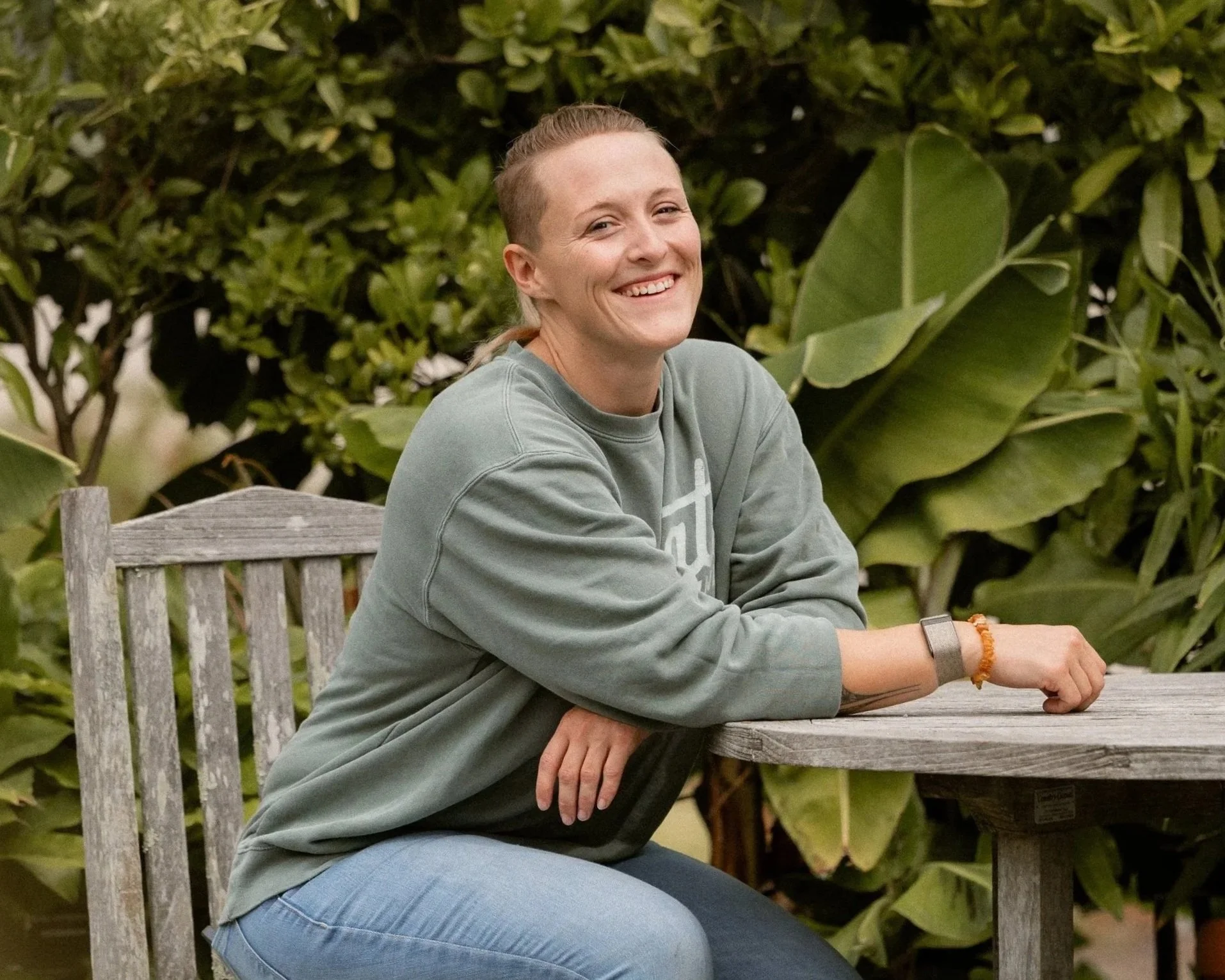 A woman with light skin and blonde hair tied back, smiling while sitting at a wooden outdoor table, wearing a gray sweatshirt and blue jeans, with greenery in the background.
