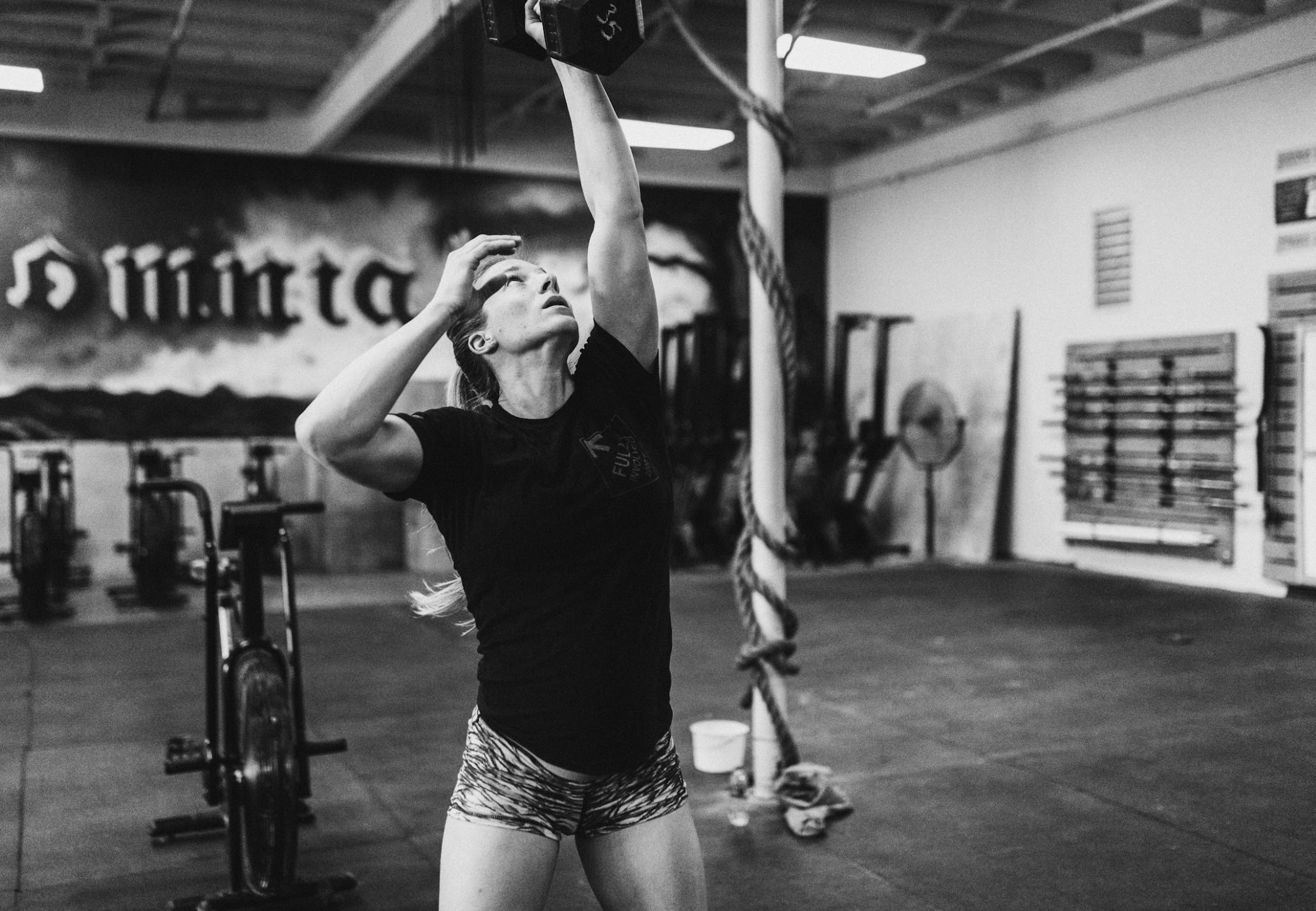 A woman in athletic wear, including a black T-shirt and patterned shorts, reaching upward with her arm extended to grab a dumbbell in a gym setting. She is standing next to a climbing rope, with gym equipment and wall-mounted fitness gear visible in 