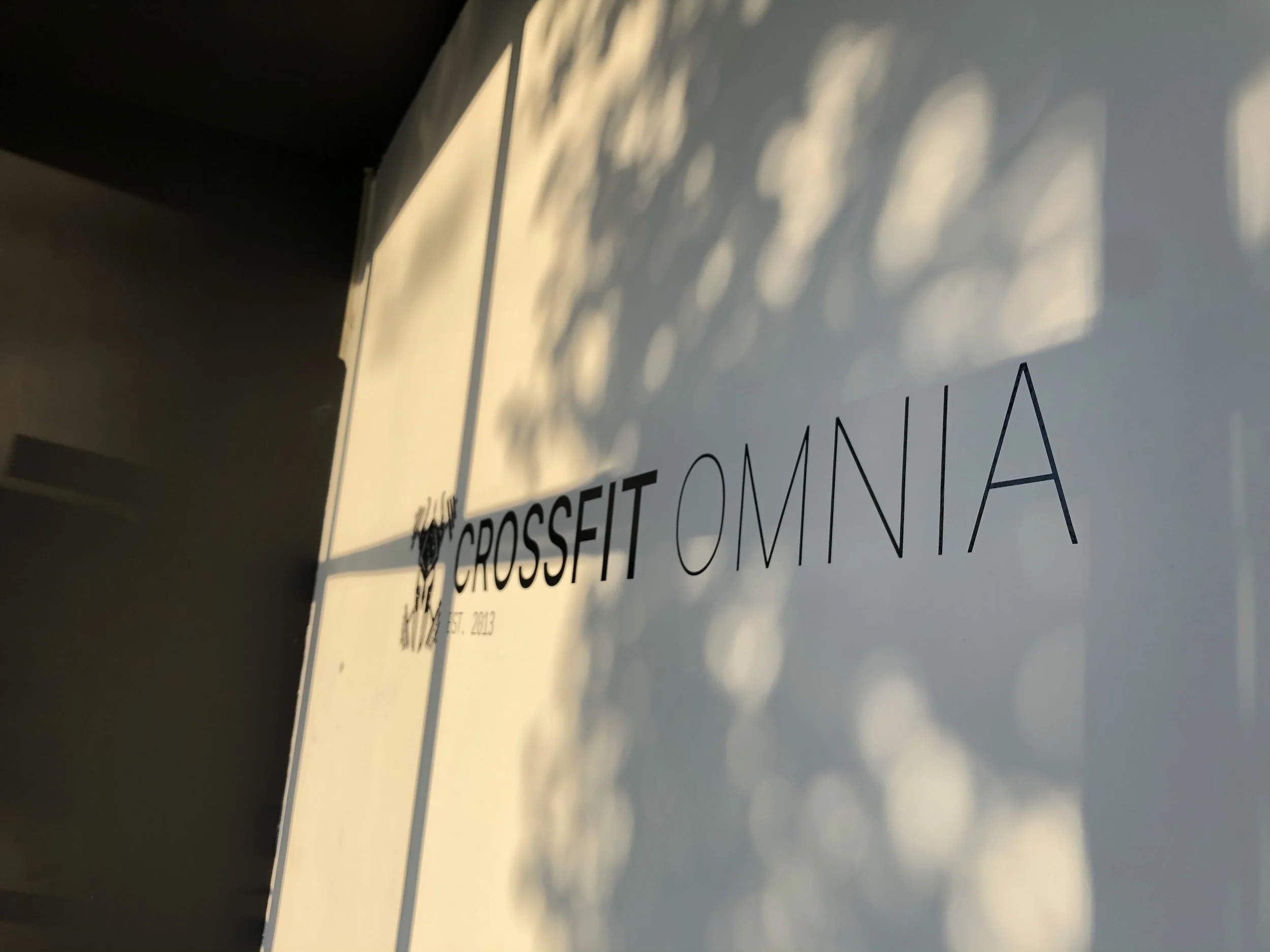 Close-up of a sign with the words 'CROSSFIT OMNIA' and a hashtag, with shadows cast on a light-colored background.