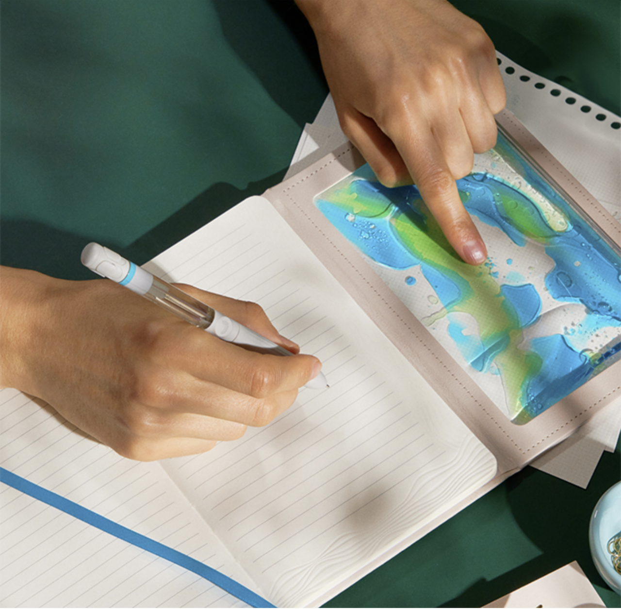 A person is writing in a notebook with a white pen, while their other hand touches a colorful, abstract, blue and green artwork in a clear plastic sleeve.