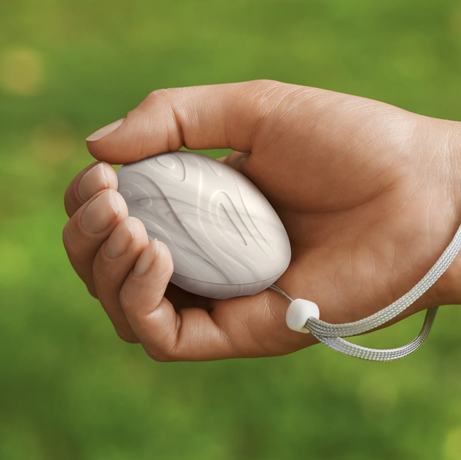 A hand holding a white, oval-shaped electronic device with a decorative pattern, attached to a lanyard, against a blurred green background.