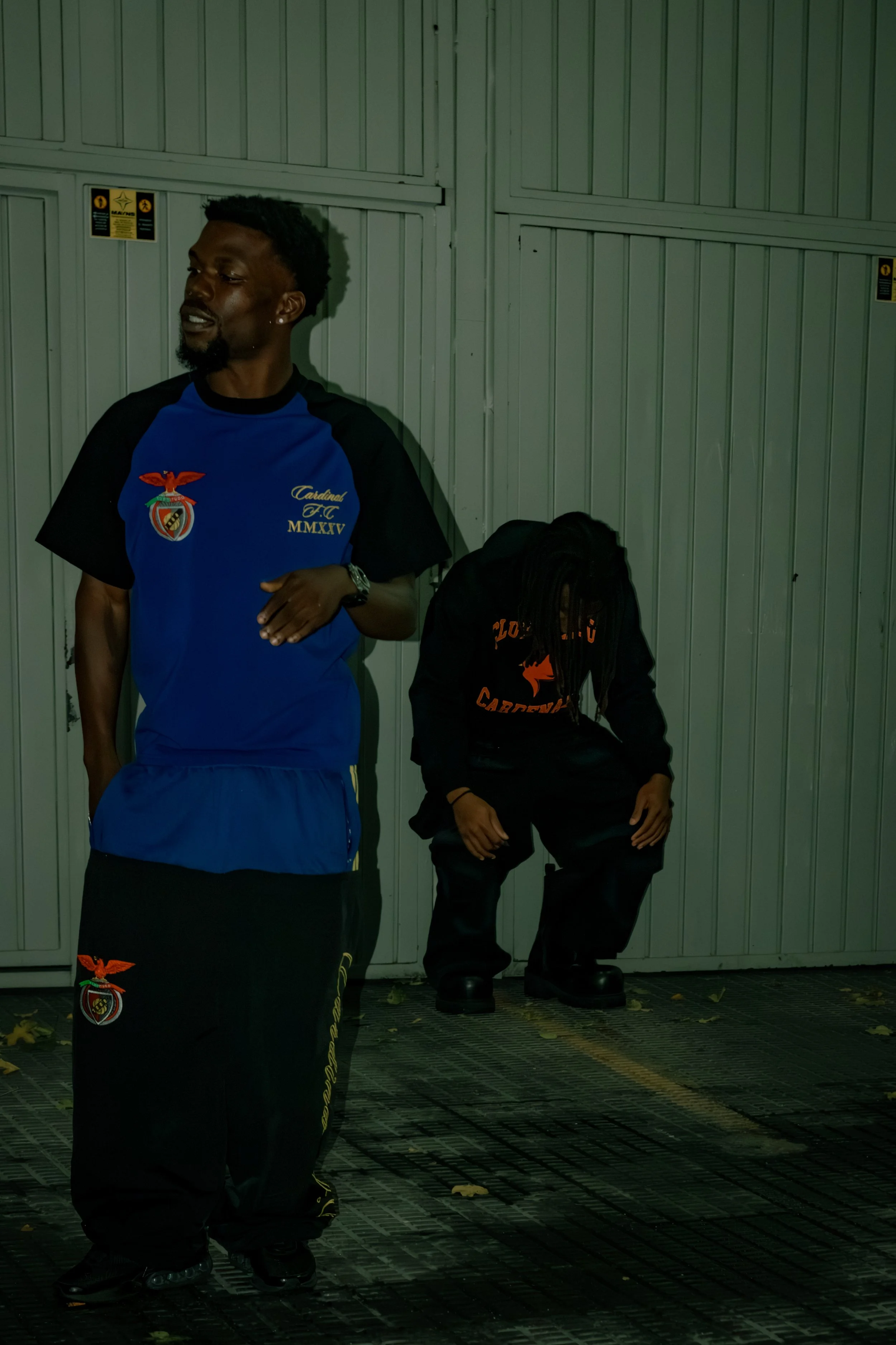 Two men standing against a gray metal wall, one in a blue sports uniform with a crest and Roman numerals, and the other dressed in black with orange lettering, sitting on the ground with their heads bowed, the man in blue smiling slightly.